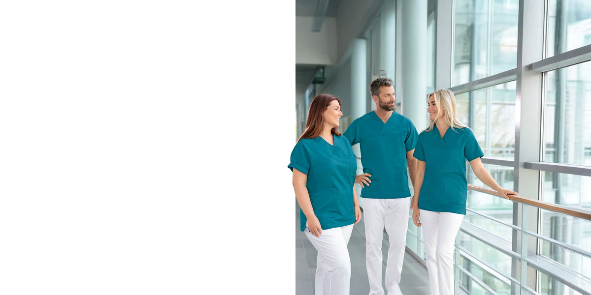 Drei medizinische Fachkräfte in türkisfarbenen OP-Kitteln und weißen Hosen stehen gemeinsam in einem hellen Krankenhausflur, blicken einander an und lächeln.