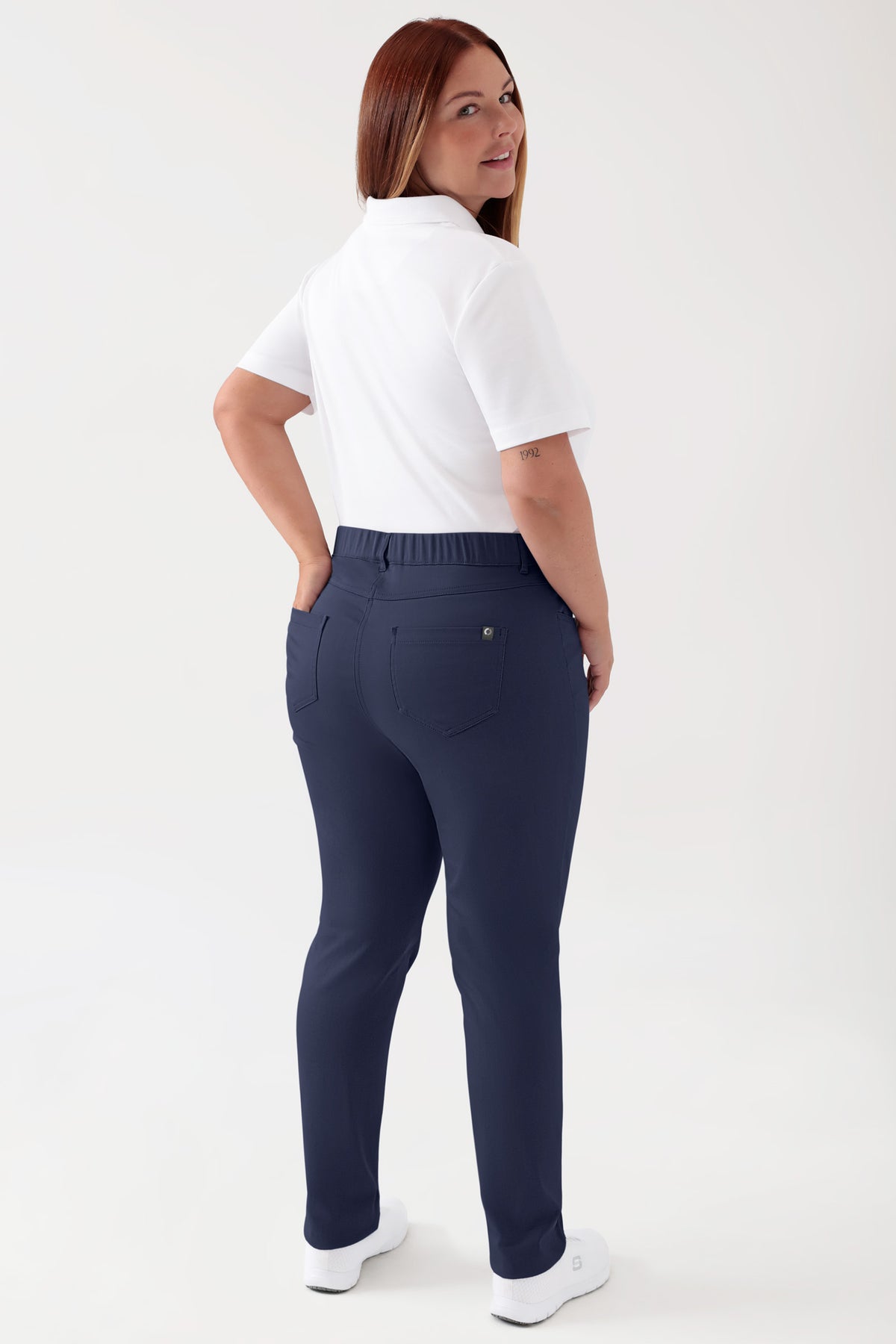 Frau in einer marineblauen COMFY FLEX 5-Pocket Hose Damen – eine Hose mit schmalem Bein in Marineblau, einem elastischen Bund mit hoher Taille und einem weißen Polohemd, die sich durch eine schlanke Silhouette auszeichnet., Rückansicht Das Model ist 173 cm groß und trägt Größe 46