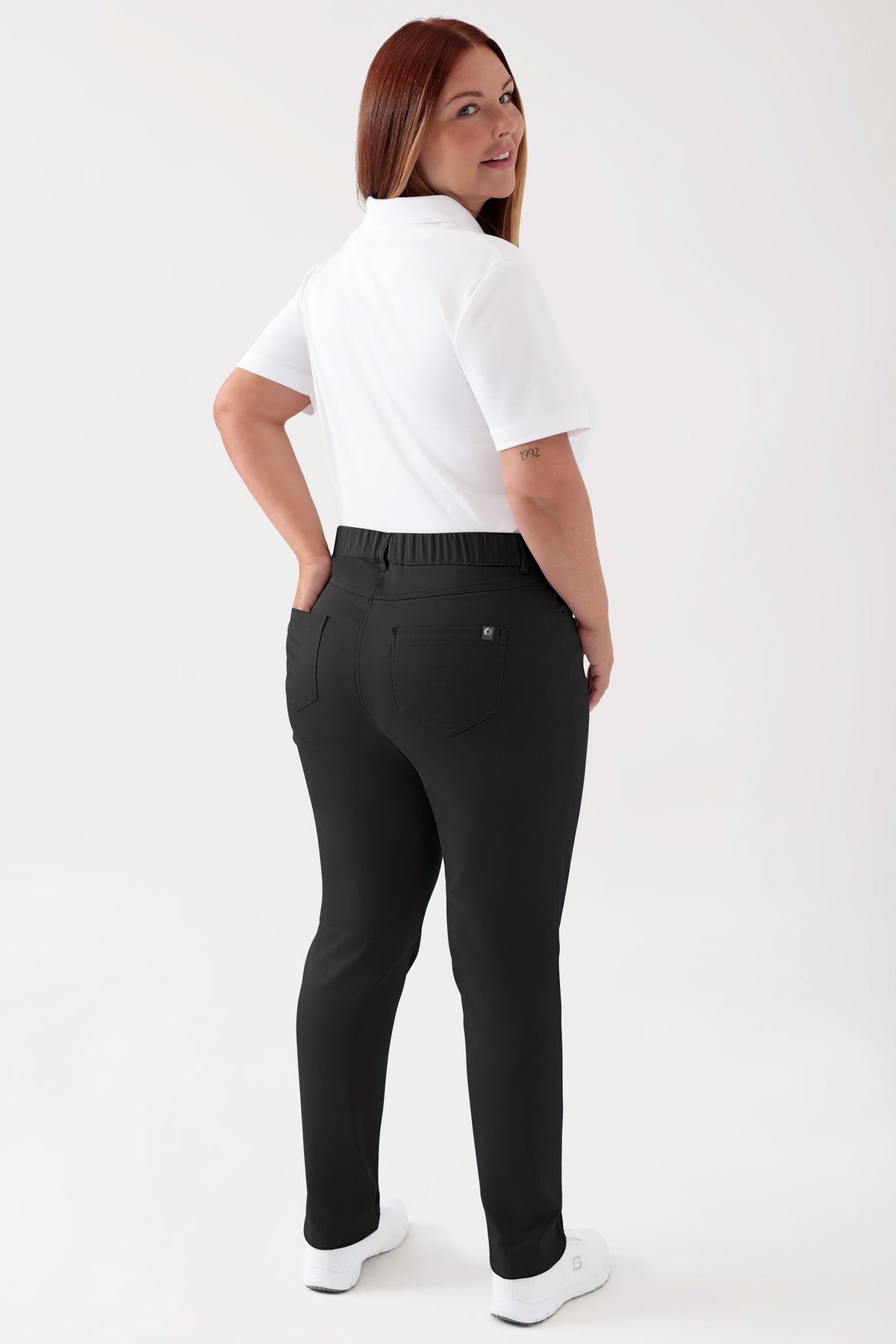 Eine Frau, die ein weißes Polohemd und eine COMFY FLEX 5-Pocket Hose Damen – schlankes Bein, schwarz trägt, die sich durch einen hohen Bund, einen elastischen Taillenbund und ein schwarzes Five-Pocket-Design in schmaler Passform auszeichnet., Rückansicht Das Model ist 173 cm groß und trägt Größe 46