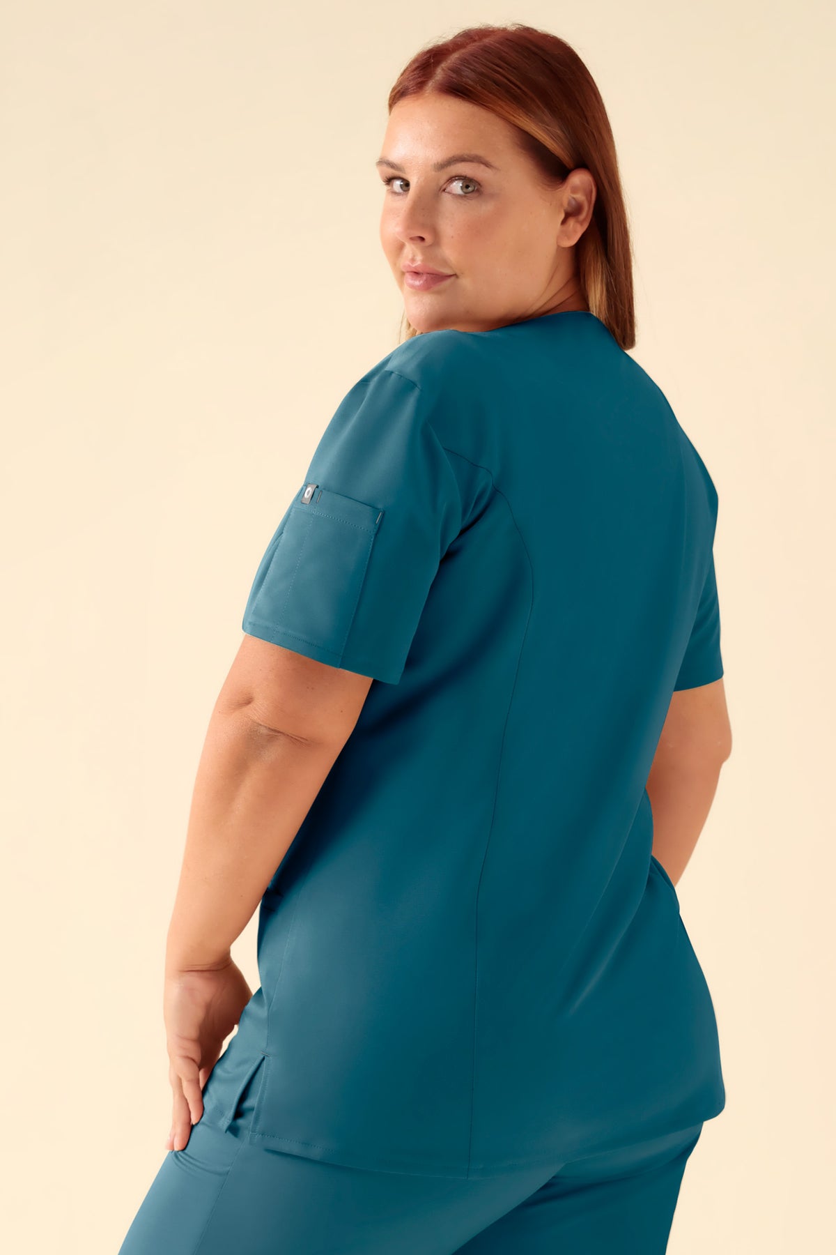 Eine Frau, die ein benzinfarbenes CLINIC DRESS KAERE Kasack Damen – durchgeknöpft trägt, ein kurzärmeliges Oberteil im medizinischen Stil mit V-Ausschnitt, Knopfleiste und einer kleinen Tasche am Ärmel., Rückansicht Das Model ist 173 cm groß und trägt Größe XL