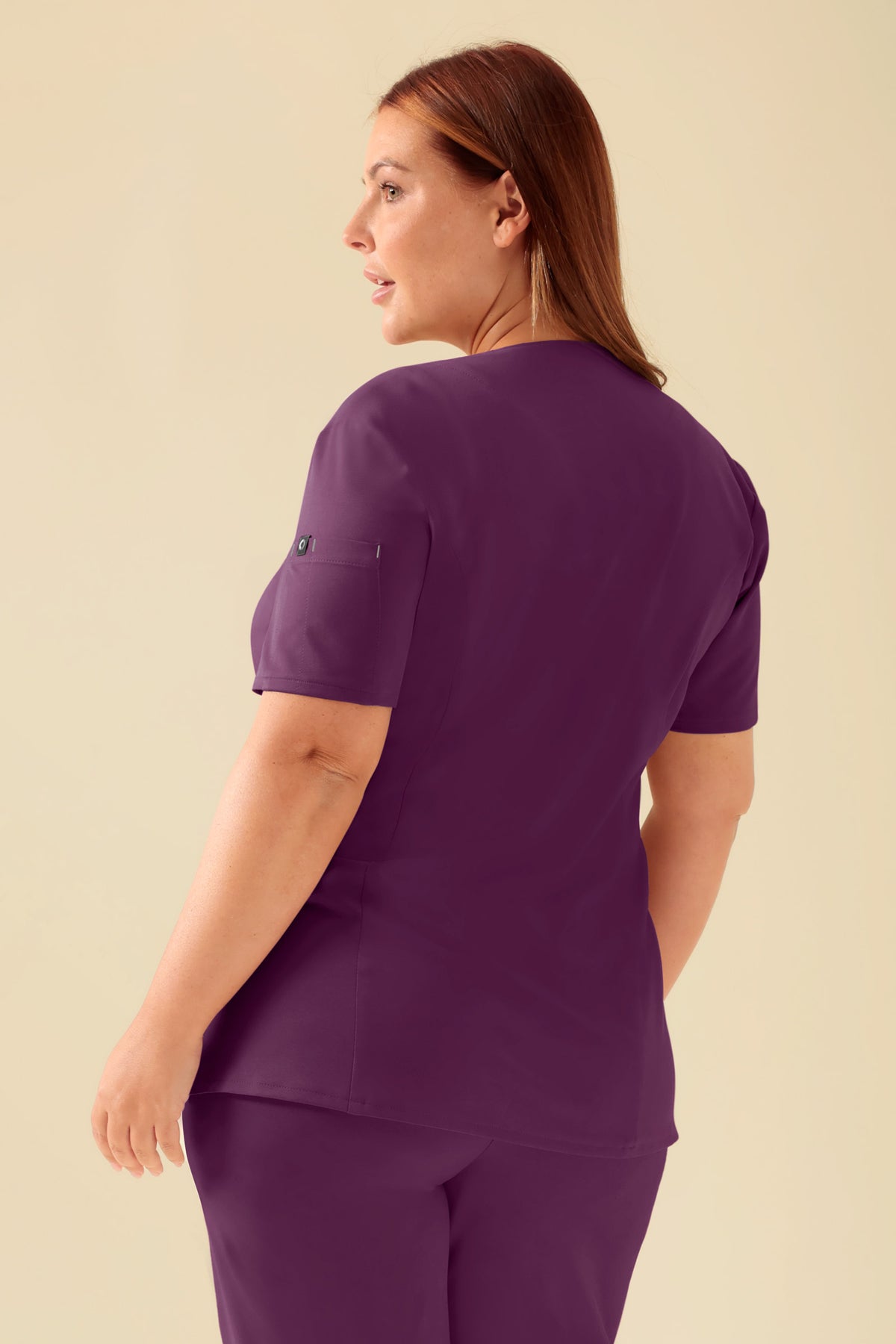 Eine Person, die ein pflaumenfarbenes KAERE Kasack Damen – durchgeknöpft pflaume Scrub-Oberteil mit V-Ausschnitt, kurzen Ärmeln und Seitentaschen trägt., Rückansicht Das Model ist 173 cm groß und trägt Größe XL