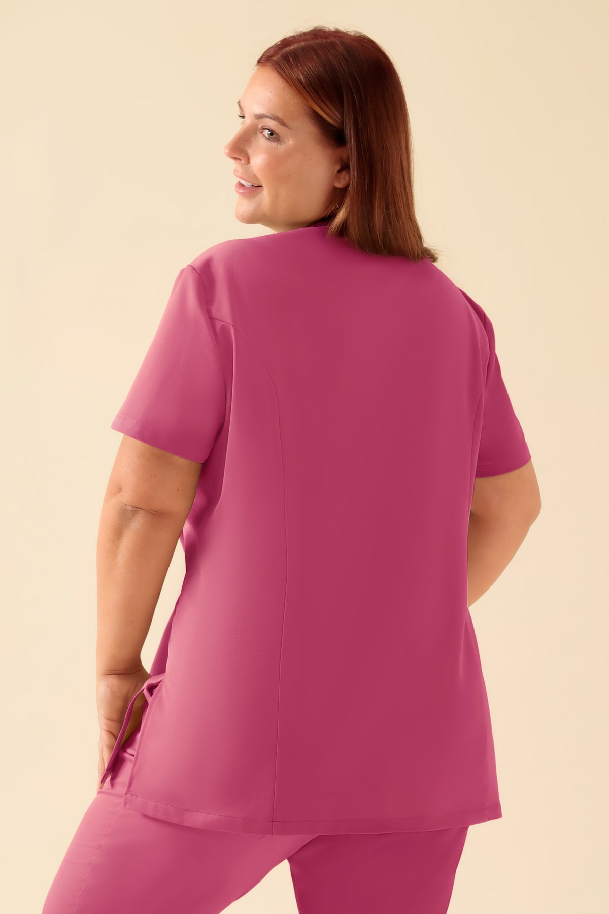 Eine Person, die einen KAERE Kasack Damen – asymmetrische Knopfleiste in Rosenholz trägt, ein tailliertes, rosafarbenes OP-Oberteil mit kurzen Ärmeln und Stehkragen., Rückansicht Das Model ist 173 cm groß und trägt Größe XL