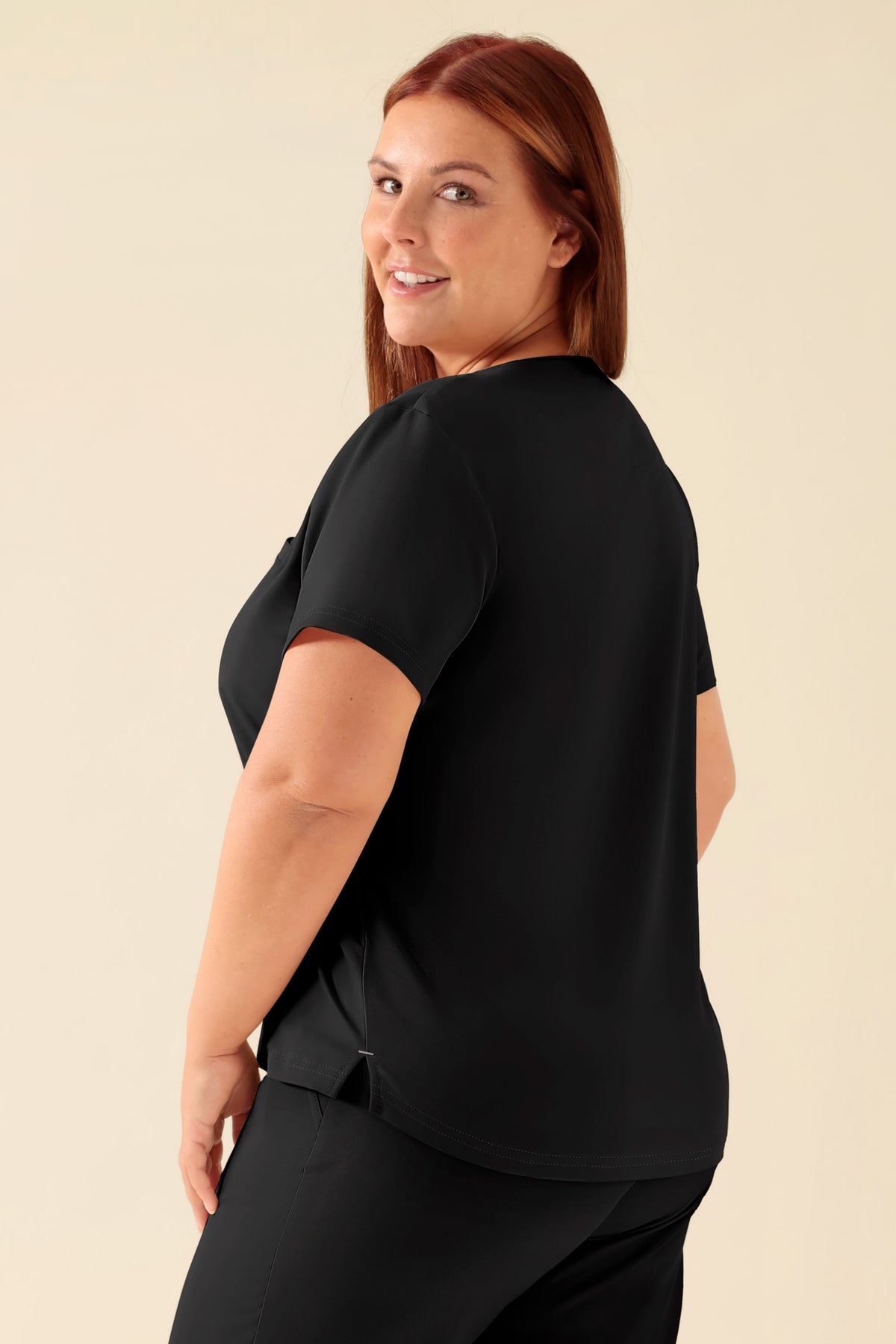 Eine Frau mit lächelndem Gesichtsausdruck blickt über ihre Schulter. Sie trägt ein schwarzes KAERE-Shirt für Damen – Rundhalsausschnitt schwarz – mit einer Brusttasche., Rückansicht Das Model ist 173 cm groß und trägt Größe XL