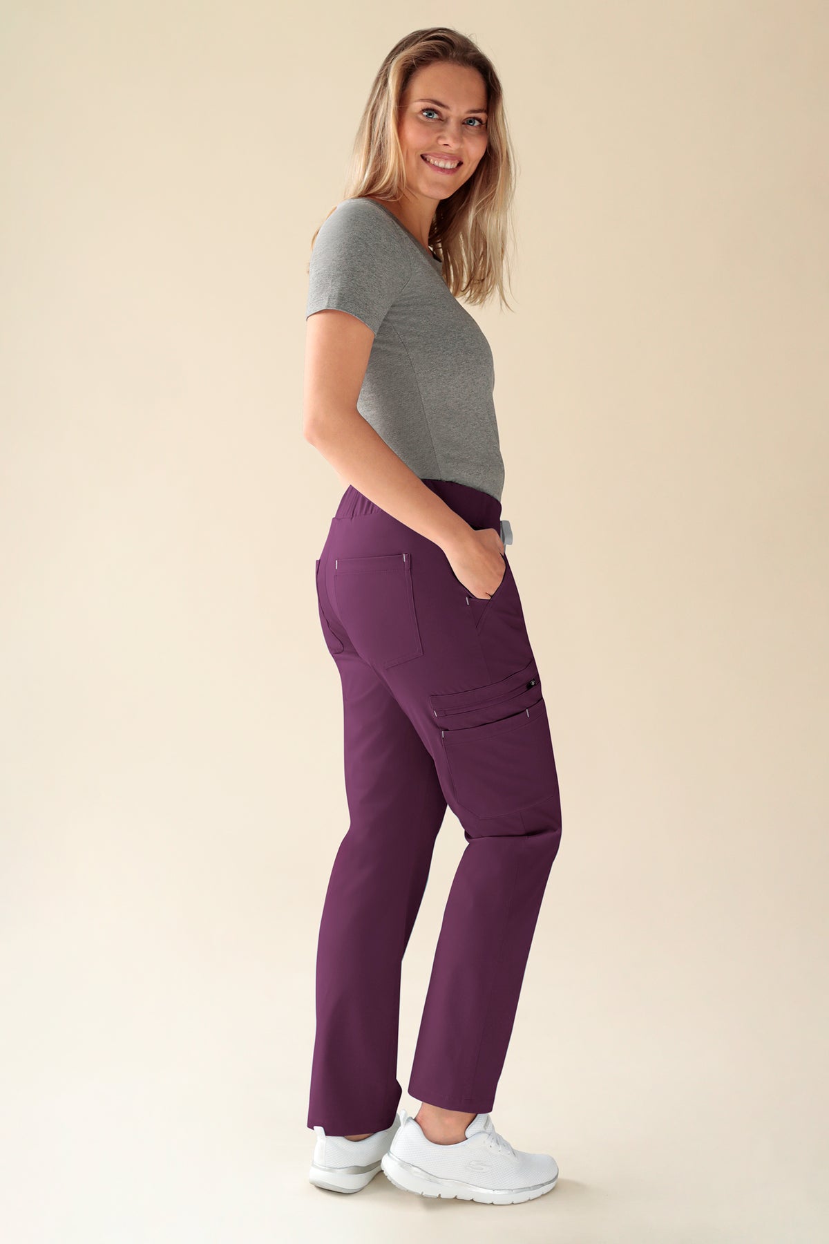 Eine Frau, die seitlich steht, trägt ein graues T-Shirt und eine pflaumenfarbene KAERE-Damenhose – gerader Saum, kurze Länge, mit Seitentaschen und kontrastierenden Riegelstichen.