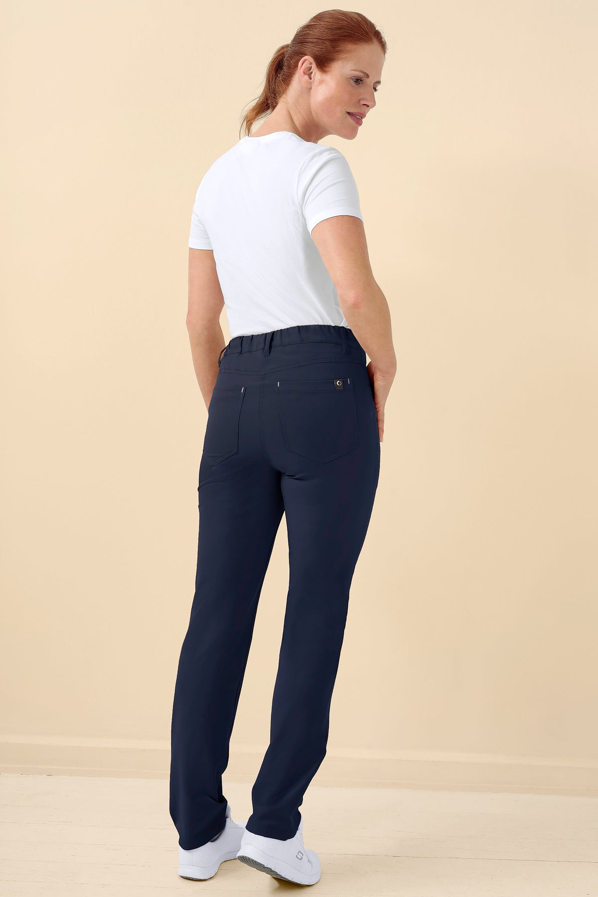 Eine Frau, die ein weißes T-Shirt und eine marineblaue KAERE Hose Damen – 5-Pocket-Hose mit bequemem Bund trägt, steht mit dem Rücken zur Kamera., Rückansicht Das Model ist 175 cm groß und trägt Größe S
