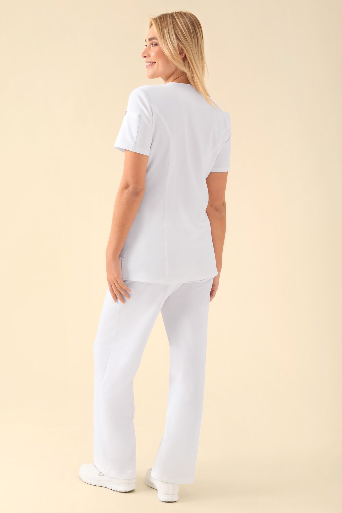 Eine Frau im Profil trägt ein weißes, durchgeknotetes KAERE Kasack Damen-Scrub-Oberteil mit Knöpfen und eine dazu passende Hose mit geradem Bein und Seitentaschen.