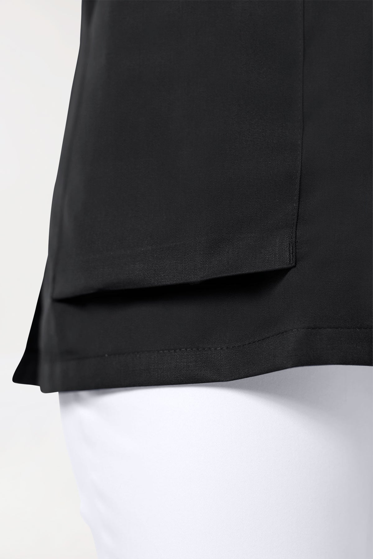 Nahaufnahme einer CORE Kasack Herren – durchgeknotete schwarze Tunika mit V-Ausschnitt, Raglanärmeln und einer großen Tasche, getragen über einer weißen Hose.