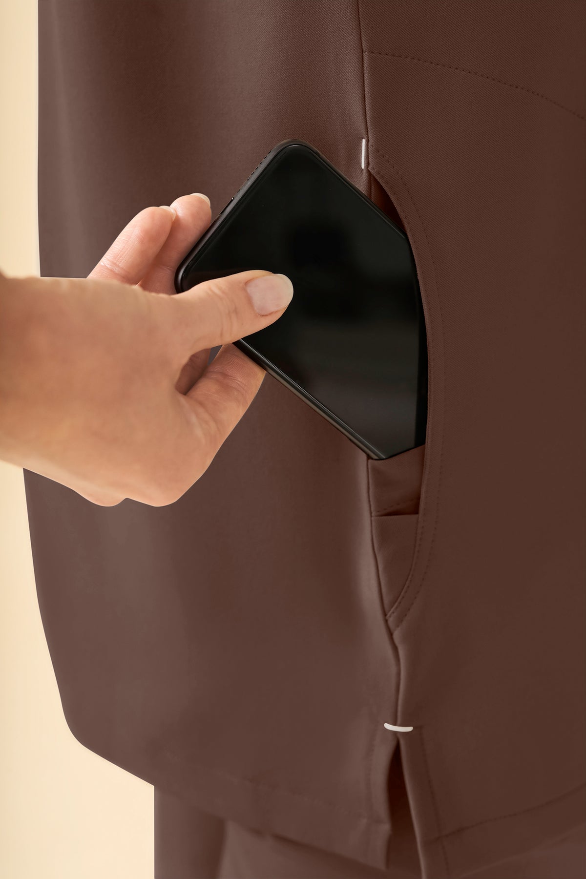 Eine Hand, die ein schwarzes Smartphone in die Seitentasche eines kaffeefarbenen KAERE Schlupfkasack Damen – Stehkragen-Tunika mit Stehkragen steckt.