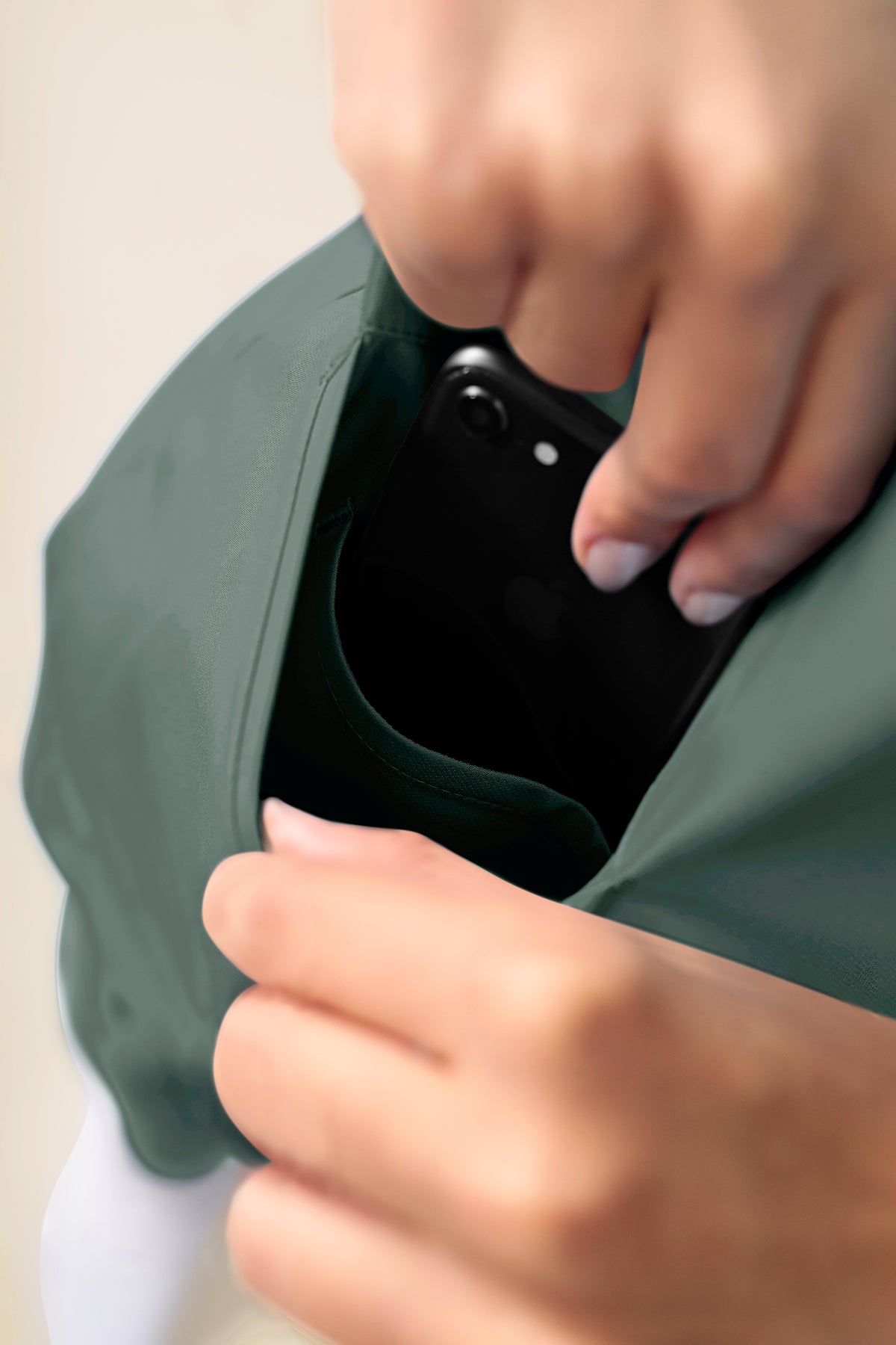 Hände, die ein schwarzes Smartphone in eine Seitentasche eines dunkelgrünen KAERE Schlupfkasack Damen – V-Ausschnitt dunkelgrün Scrub-Oberteils stecken.