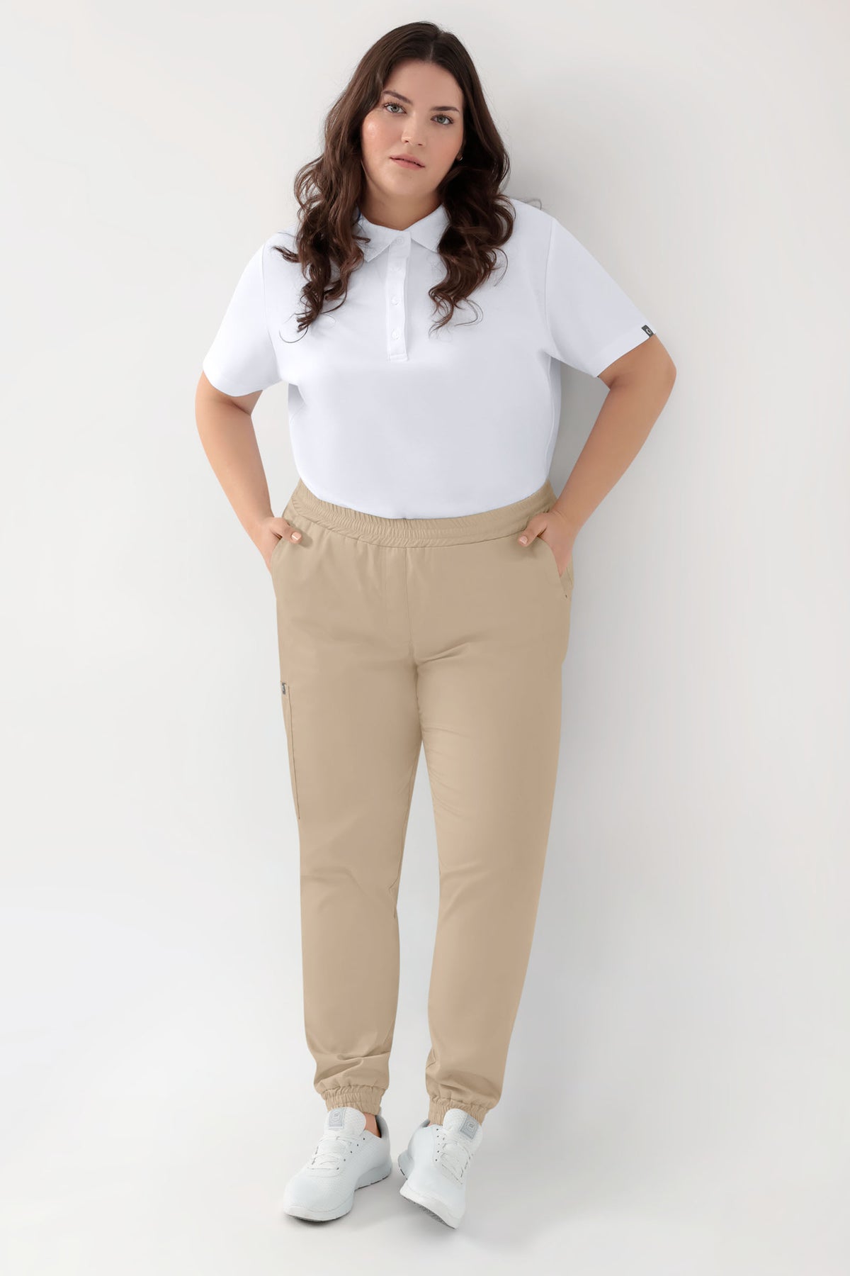 Eine Person, die ein weißes Poloshirt, eine GRÜNE Stretchhose für Damen – im Jogginghosen-Stil, sandfarben, mit Rundum-Gummibund und lockerer Beinweite – sowie weiße Sneaker trägt.