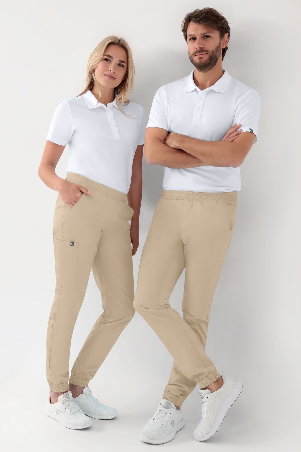Eine Frau und ein Mann in weißen Poloshirts und sandfarbenen GREEN Stretch-Strumpfhosen für Damen – im Jogginghosen-Stil, sandfarben, mit elastischem Bund und lockerem Bein, dazu weiße Sportschuhe.