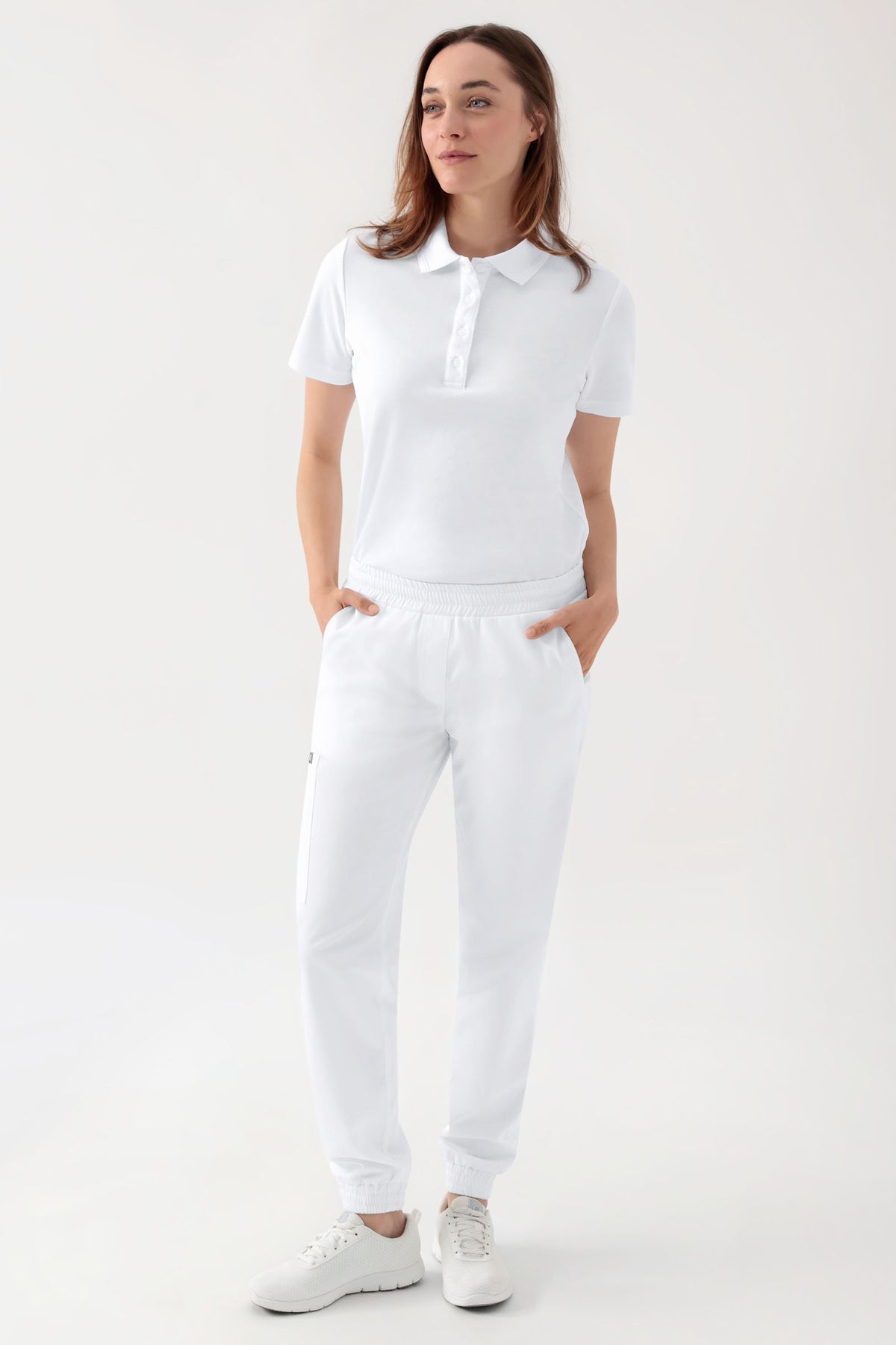 Frau in einem weißen, kurzärmeligen Polohemd, einer weißen GREEN Stretch-Hose für Damen – im Jogginghosen-Stil, weiß, mit bequemem elastischem Bund und lockerer Passform an den Beinen – sowie weißen Sneakers.