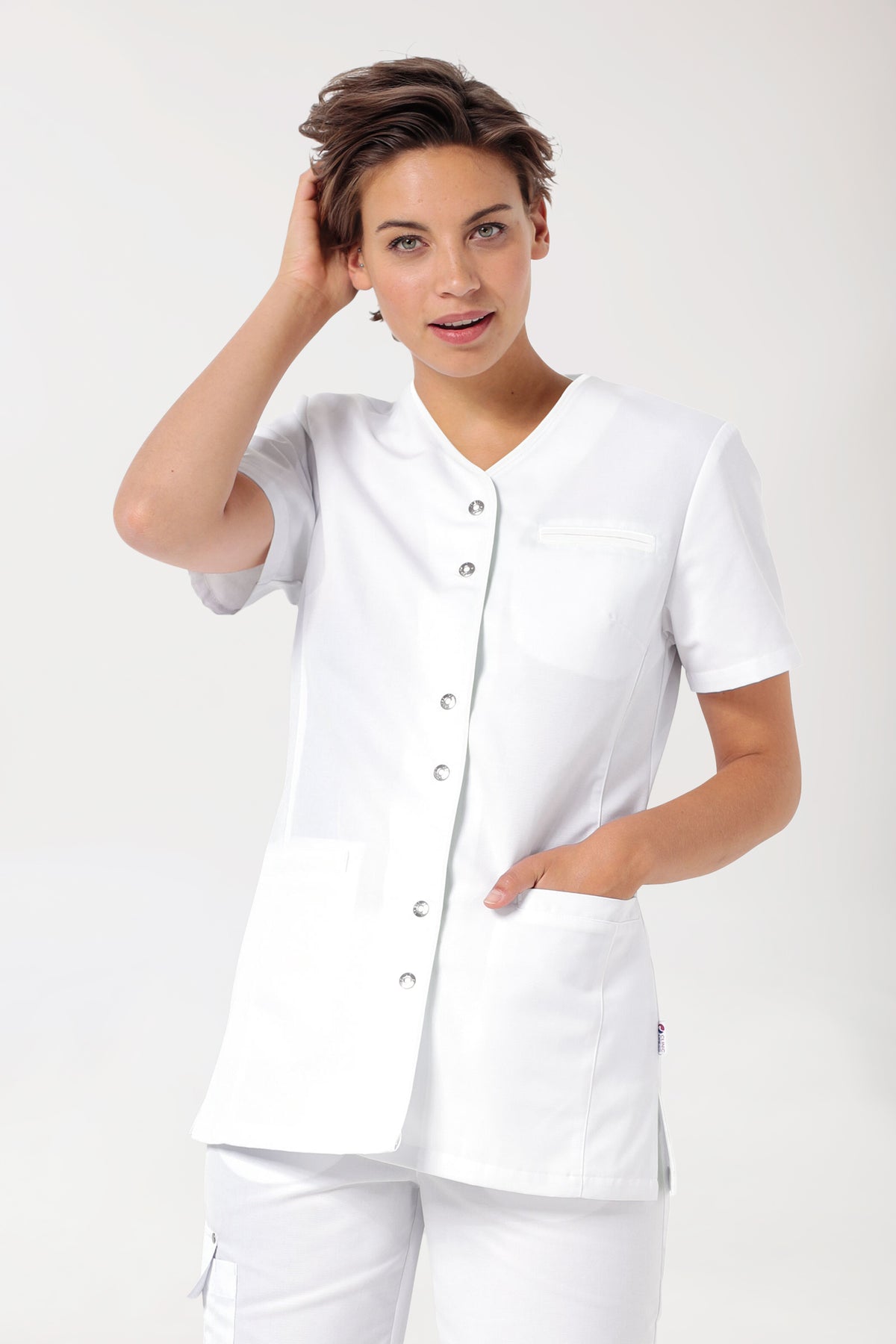 Eine Person, die ein weißes CLINIC DRESS Beauty Kasack Damen – Druckknopfleiste trägt, eine kurzärmelige Arztkittel mit V-Ausschnitt, Druckknopfleiste vorne und Seitentaschen.