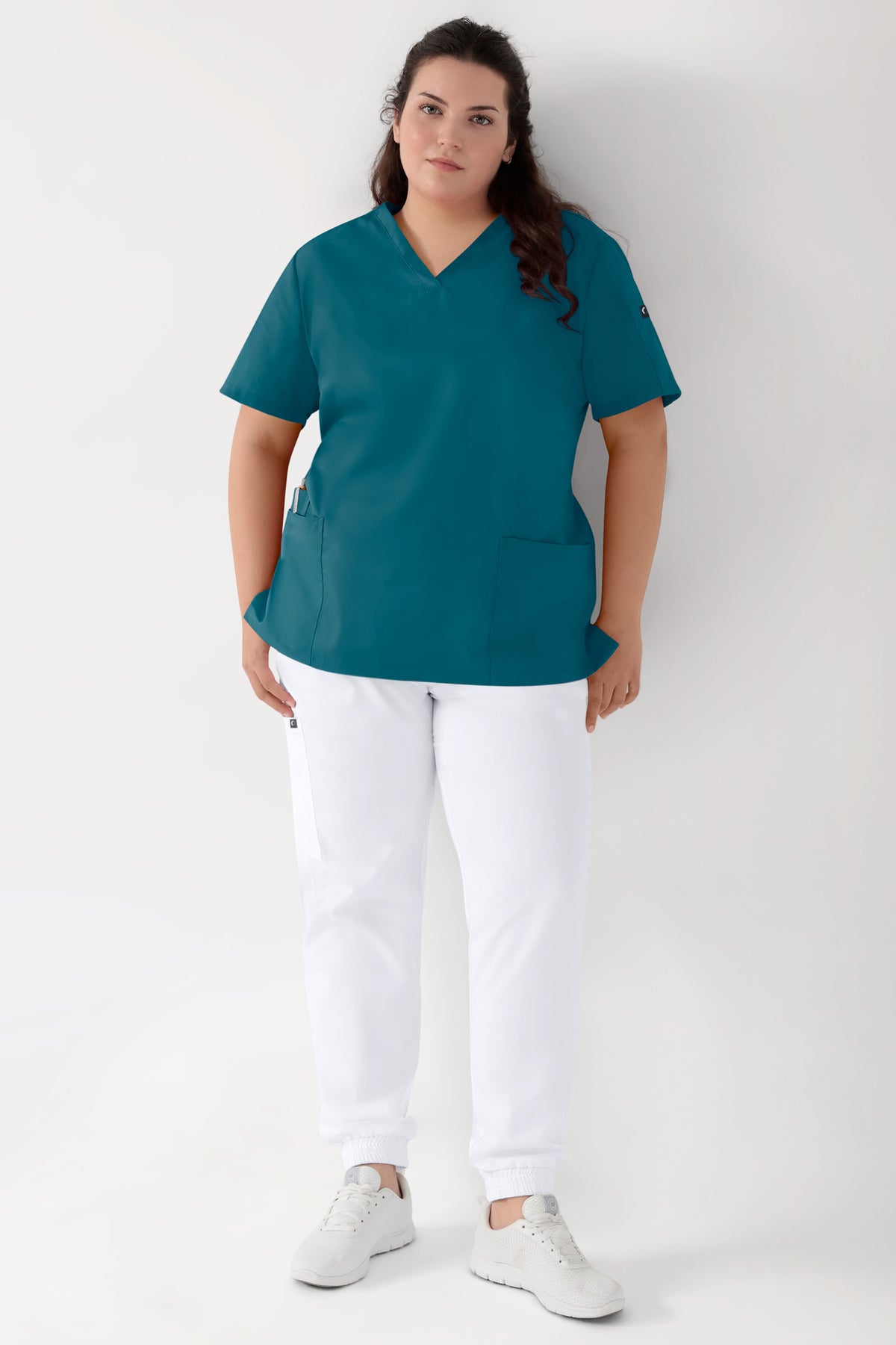 Eine Person, die einen GRÜNEN Stretch-Schlupfkasack für Damen – V-Ausschnitt in Petrol-Teal – sowie eine weiße Jogginghose im Scrub-Stil mit Seitentaschen und weiße Sneaker trägt.
