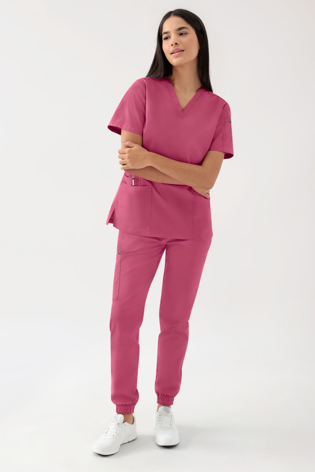 Eine Frau, die ein grünes Stretch-Schlupfkleid von CLINIC DRESS GREEN für Damen – mit V-Ausschnitt und Rosenholz-Muster – sowie eine dazu passende Jogginghose mit Seitentaschen und weiße Sneaker trägt.