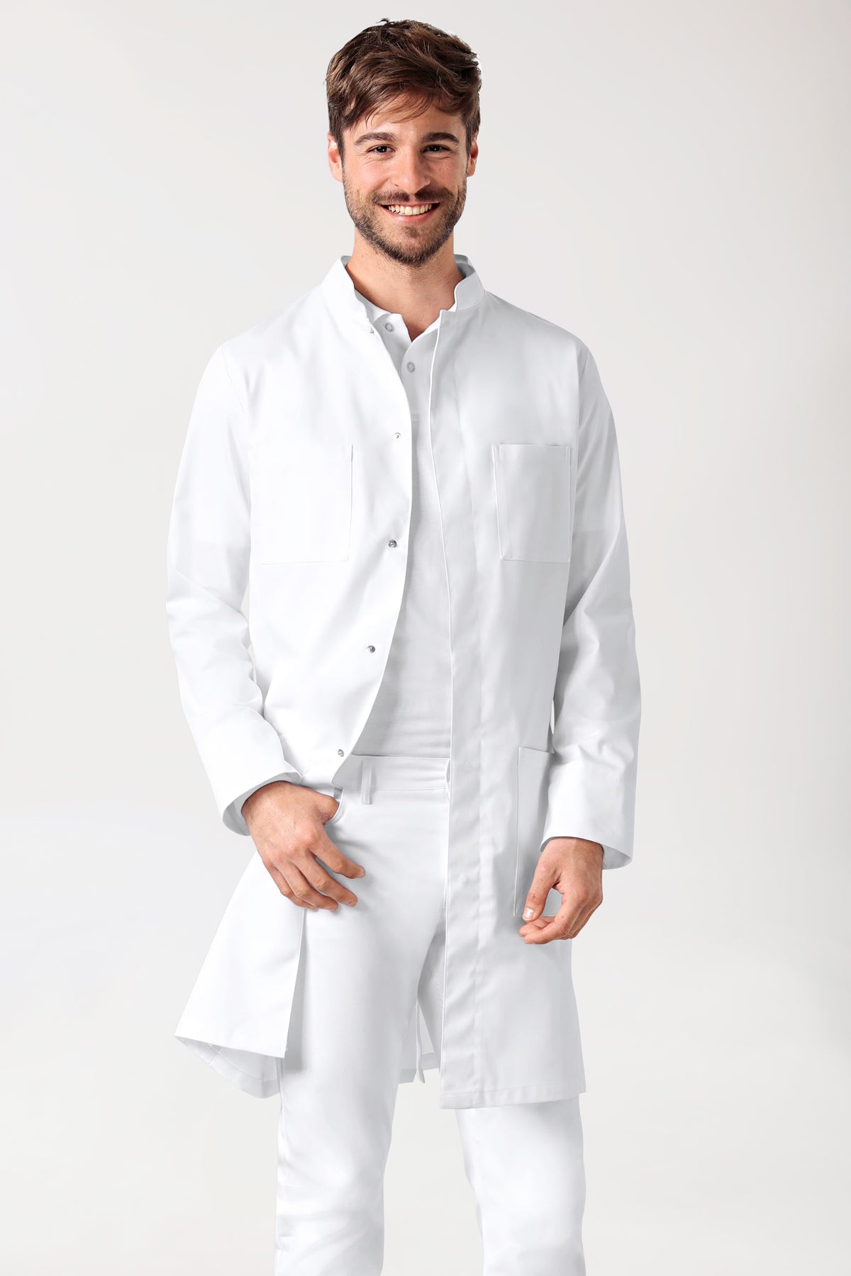 Ein lächelnder Mann trägt einen weißen CLINIC DRESS Mantel Herren – Druckknopfleiste – Stehkragen weiß mit Stehkragen und verdeckter Druckknopfleiste über einem weißen Hemd und einer weißen Hose.