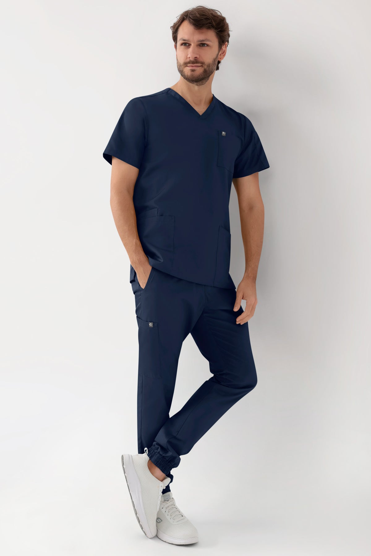 Ein Mann trägt einen marineblauen GREEN Stretch-Schlupfkasack für Herren – V-Ausschnitt in Marineblau mit einer Armtasche, dazu eine passende Jogginghose mit Seitentaschen und weiße Sneaker.