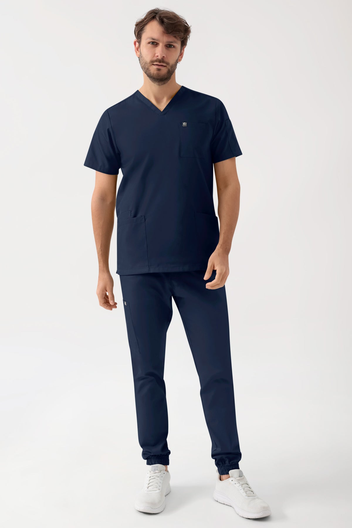 Ein Mann, der ein marinegrünes, elastisches Herren-Schlupfhemd mit V-Ausschnitt und einer Armtasche, eine dazu passende Jogginghose mit Seitentaschen sowie weiße Sportschuhe trägt.