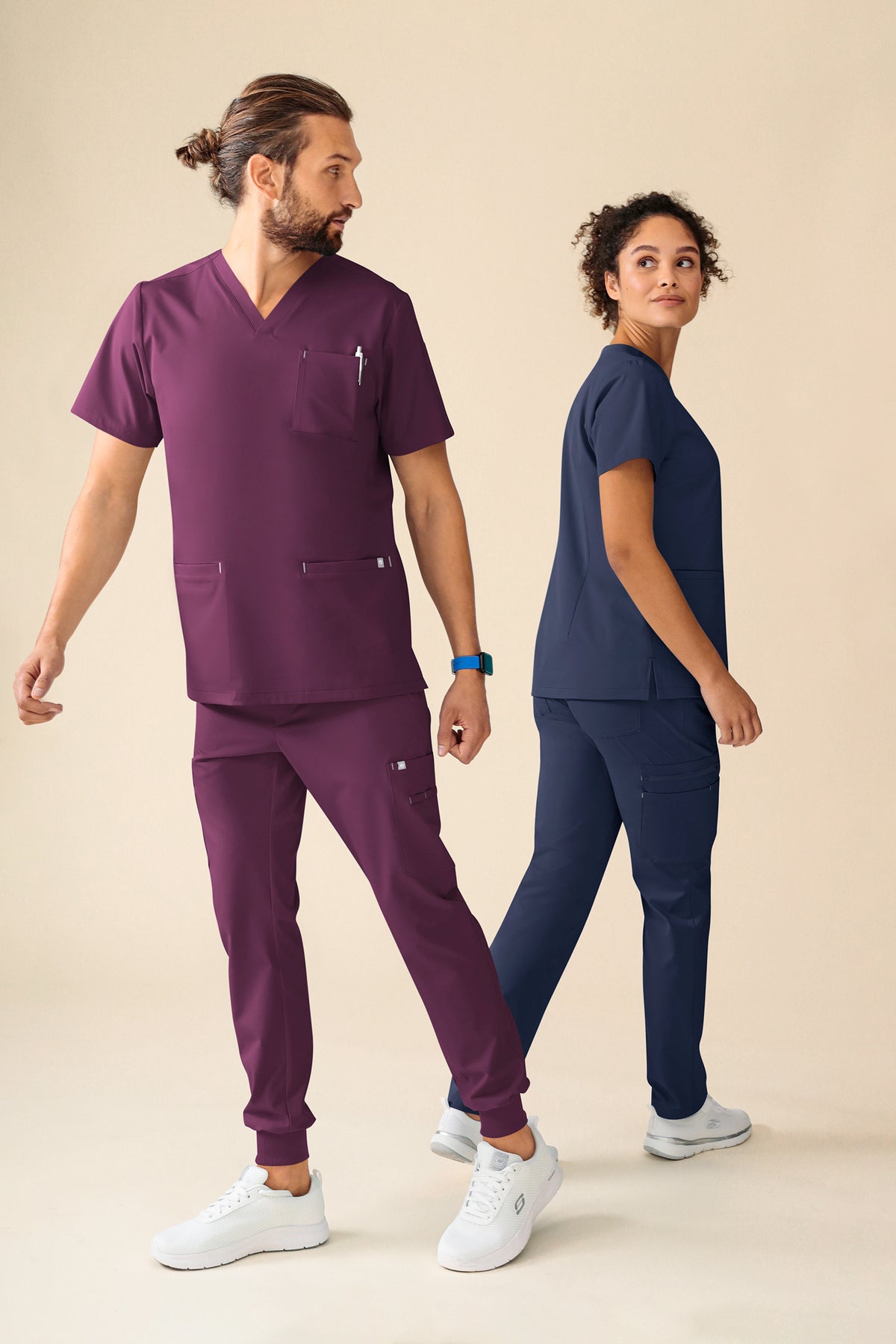 Ein Mann, der ein KAERE Schlupfkasack Herren – V-Ausschnitt pflaume Scrub-Oberteil und eine dazu passende Jogginghose trägt, sowie eine Frau, die ein marineblaues Scrub-Oberteil mit V-Ausschnitt und eine Hose trägt.