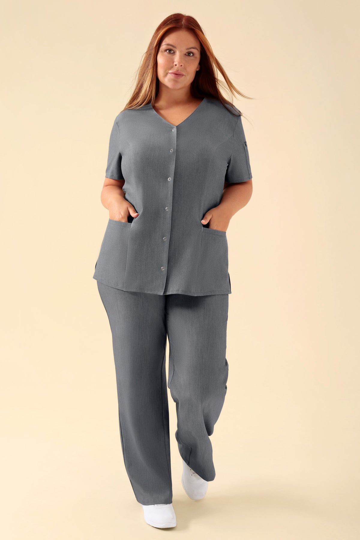 Eine Frau, die einen dunkelgrau melierten KAERE Kasack Damen von CLINIC DRESS trägt, eine Tunika mit V-Ausschnitt, Knopfleiste vorne, Seitentaschen und einer dazu passenden Hose., Vorderansicht Das Model ist 173 cm groß und trägt Größe XL