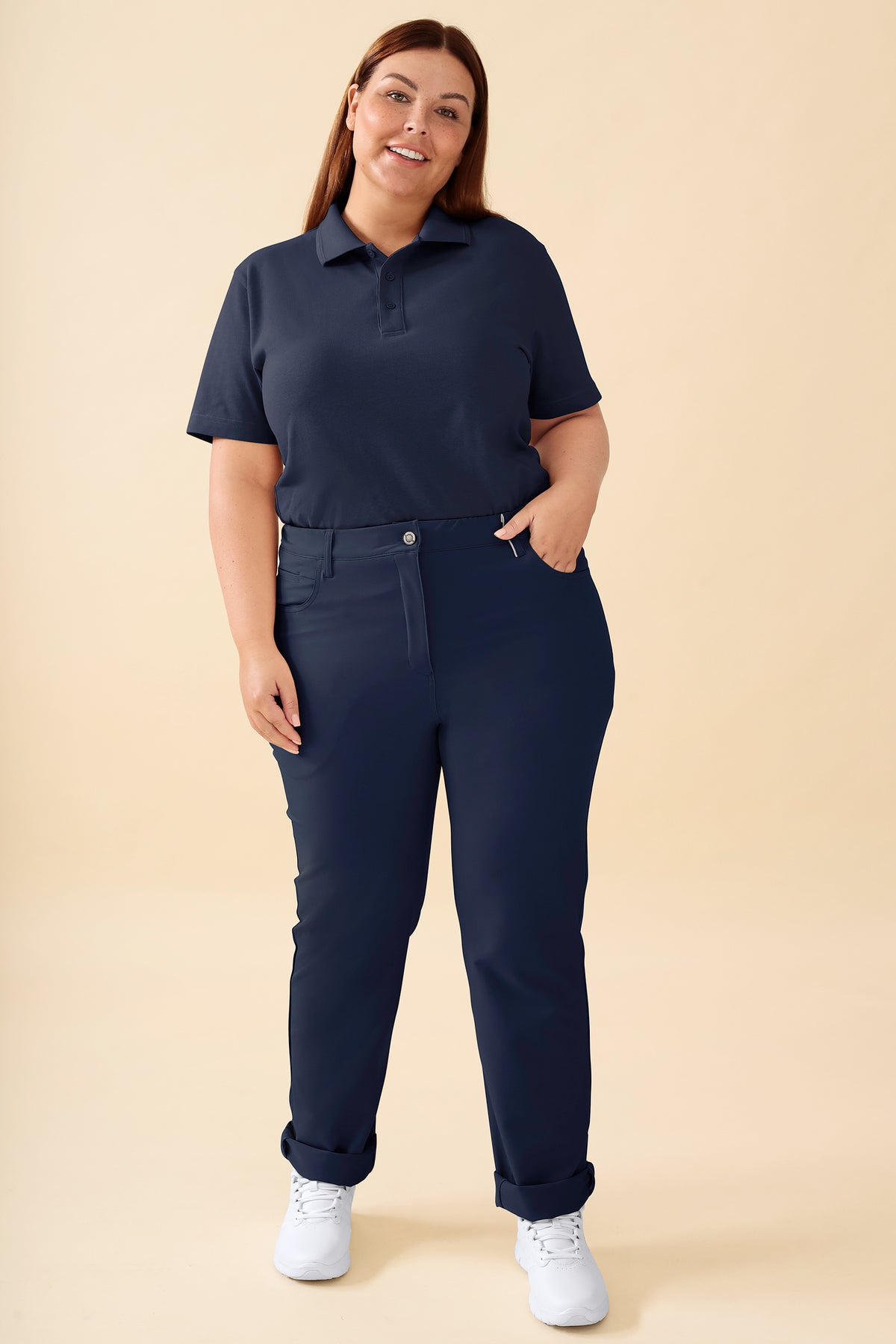 Eine Frau, die ein marineblaues Poloshirt, eine Hose von CLINIC DRESS KAERE – eine marineblaue 5-Pocket-Hose mit Umschlag am Saum – sowie weiße Sneaker trägt., Vorderansicht Das Model ist 173 cm groß und trägt Größe XL