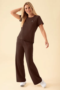 Eine lächelnde Frau, die ein kaffeefarbenes KAERE-Shirt für Damen mit Rundhalsausschnitt und Brusttasche trägt, dazu eine passende weit geschnittene OP-Hose und weiße Sneaker.