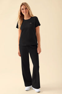 Eine Frau, die ein schwarzes KAERE-Shirt für Damen – Rundhalsausschnitt, schwarz – mit Rundhalsausschnitt und Brusttasche trägt, dazu eine passende schwarze Hose.