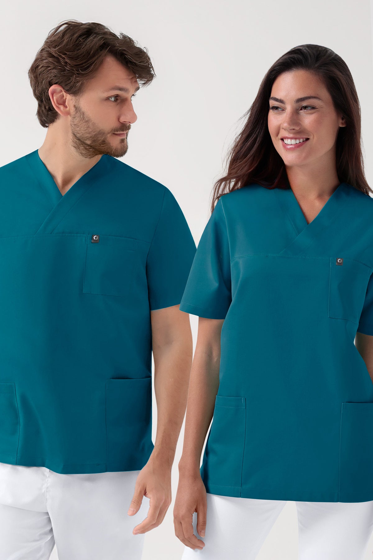 Ein Mann und eine Frau, die GRÜNE Stretch-Schlupfkasack-Unisex-Obershirts mit V-Ausschnitt in der Farbe Petrol und Seitentaschen sowie weiße Hosen tragen.
