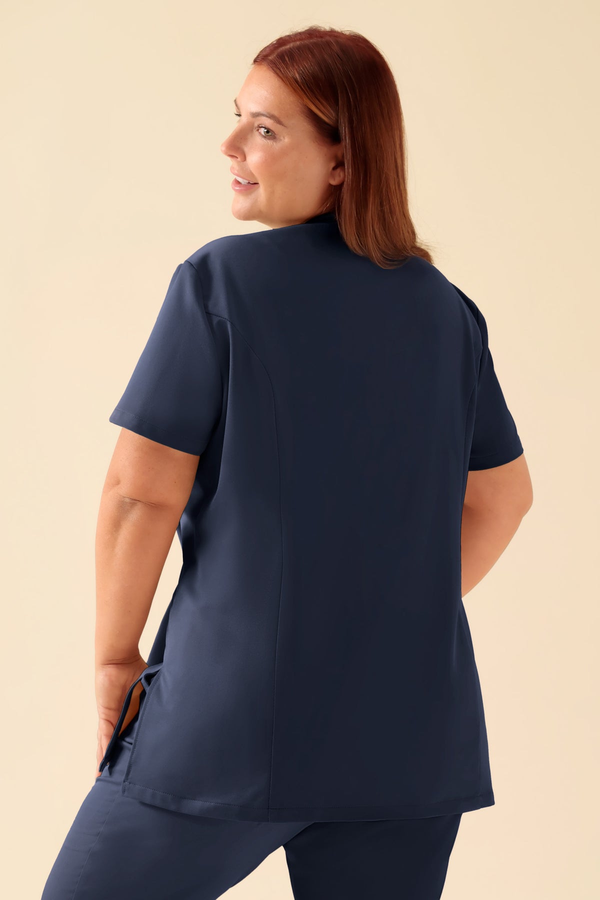 Eine Person, von hinten gesehen, die ein KAERE Kasack Damen – asymmetrische Knopfleiste, marineblaues Scrub-Oberteil mit tailliertem, marineblauem Schnitt und Stehkragen trägt., Rückansicht Das Model ist 173 cm groß und trägt Größe XL
