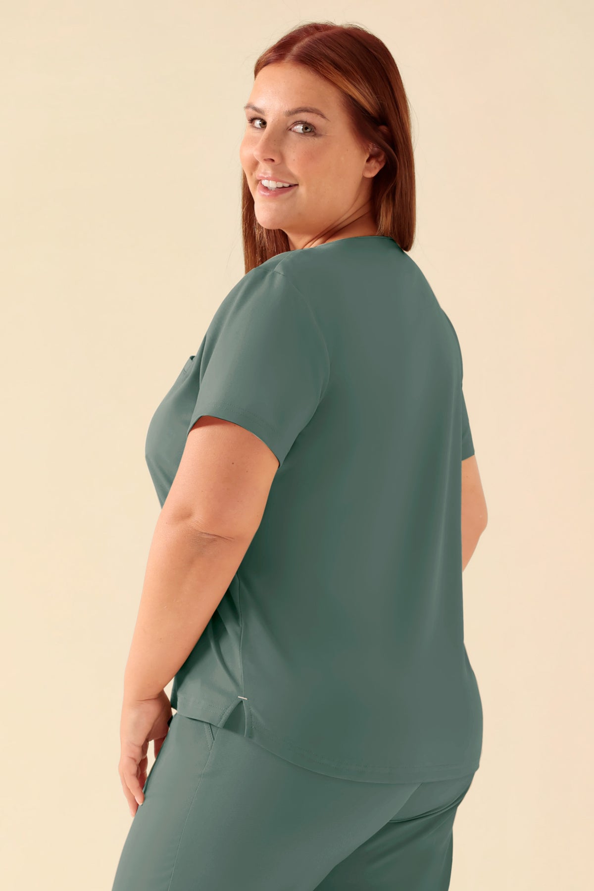 Eine Person, die ein KAERE-Shirt Damen – Rundhalsausschnitt dunkelgrün trägt, ein kurzärmeliges, dunkelgrünes OP-Oberteil mit Rundhalsausschnitt und einer Brusttasche., Rückansicht Das Model ist 173 cm groß und trägt Größe XL