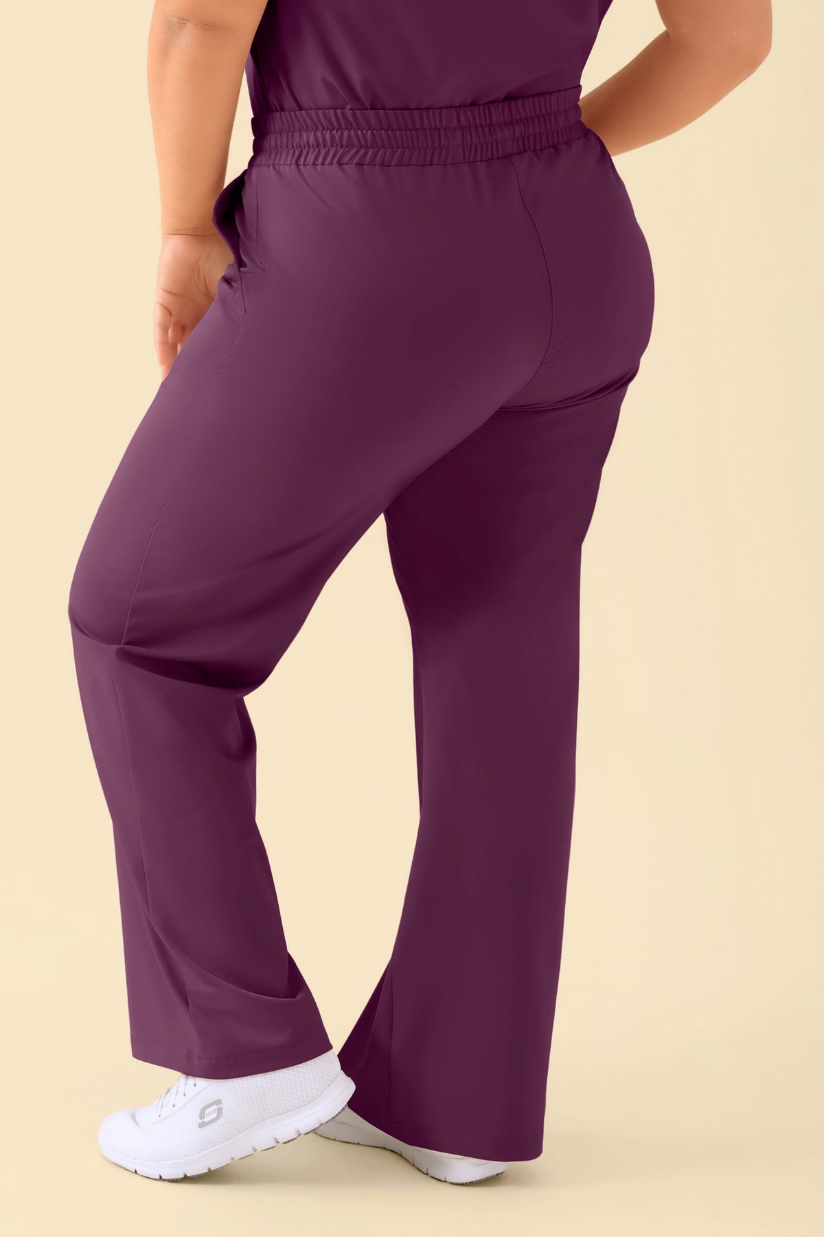 Eine Person trägt eine pflaumenfarbene KAERE-Hose Damen – weites Bein, Pflaume – eine Scrub-Hose mit lässigem, weitem Schnitt und einem bequemen elastischen Bund, kombiniert mit weißen Sneakers., Rückansicht 