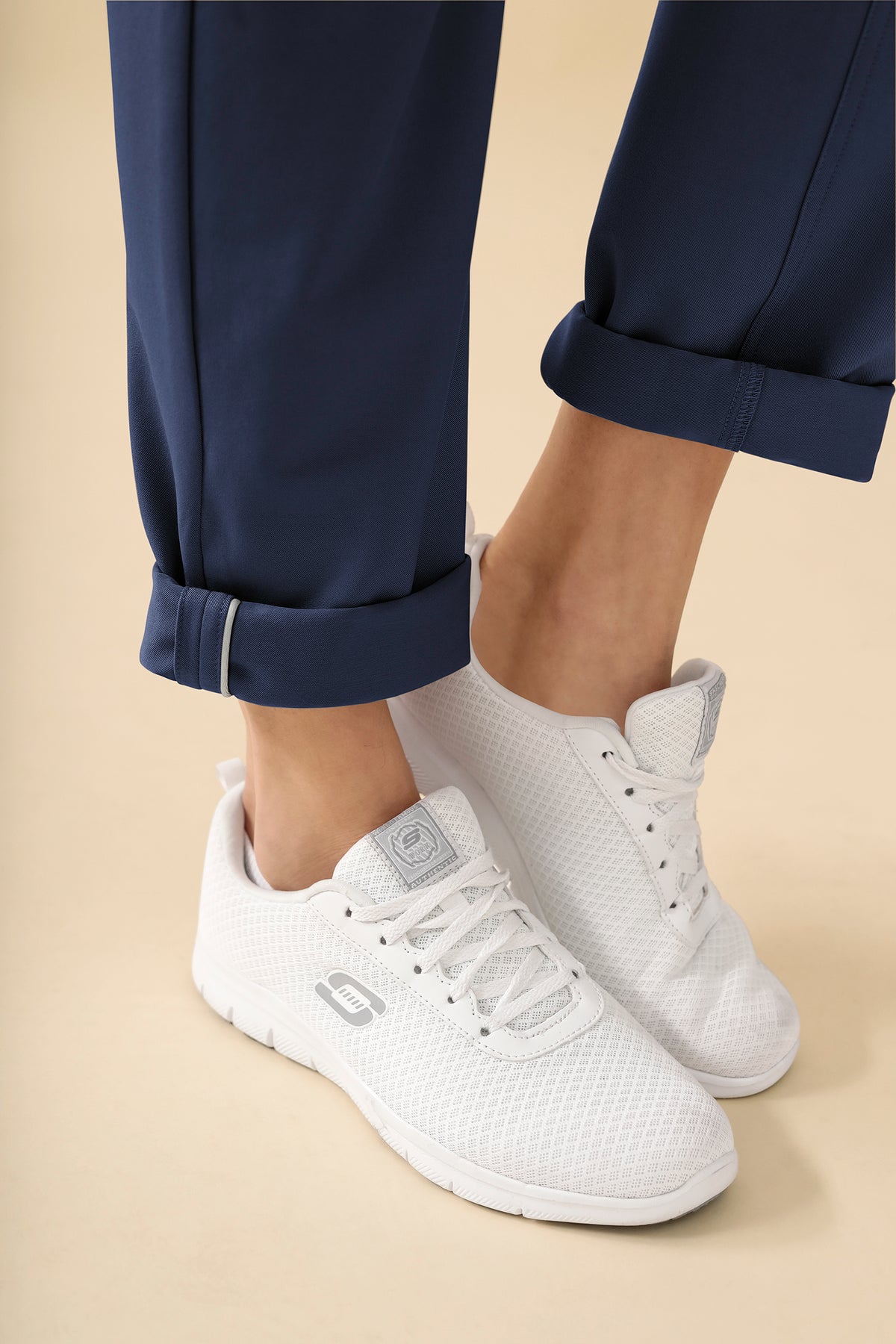 Nahaufnahme einer Person, die eine KAERE Hose Damen – eine marineblaue 5-Pocket-Hose mit Umschlägen – und weiße Schnürsneaker trägt.