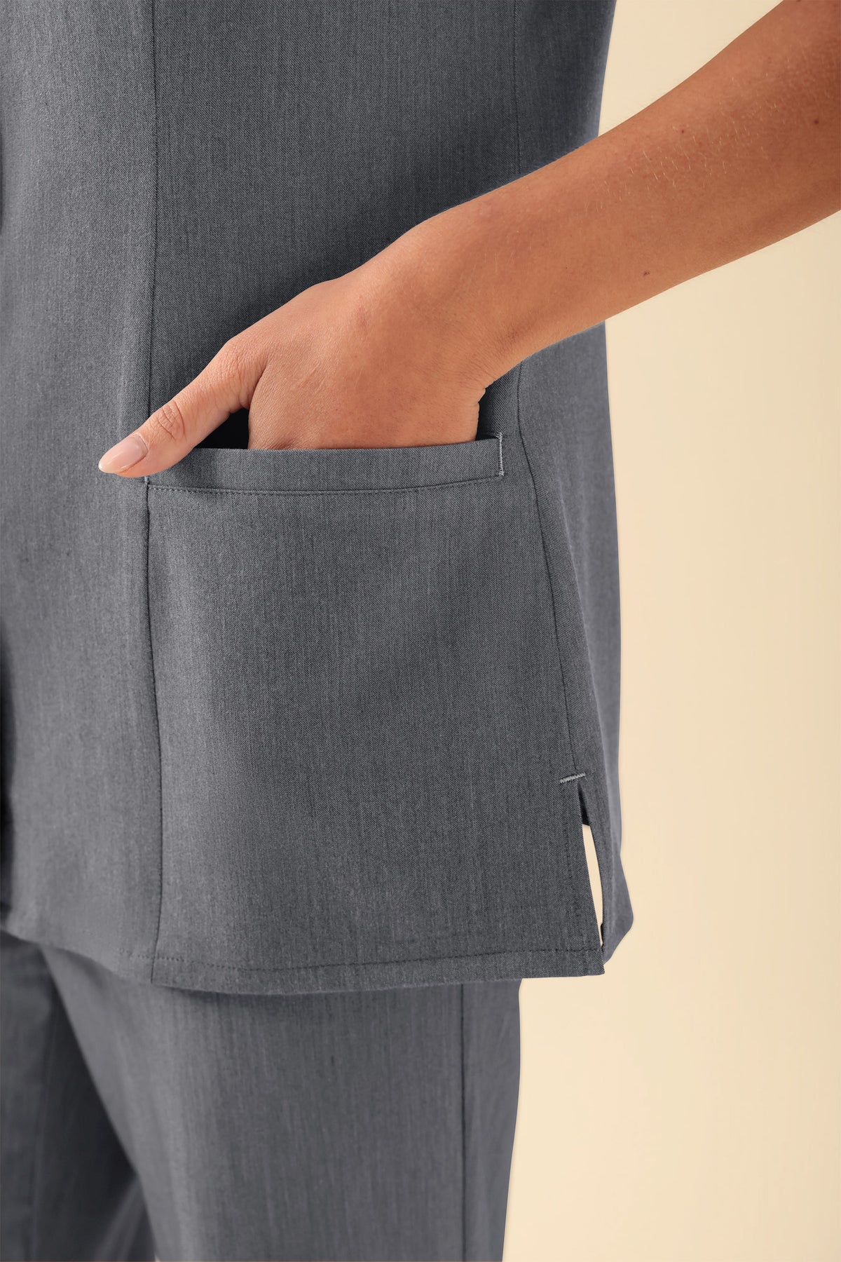 Eine Hand ruht in der Seitentasche eines dunkelgrauen, melierten KAERE Kasack Damen von CLINIC DRESS, eines Scrub-Oberteils mit V-Ausschnitt und einem Schlitz am Saum.