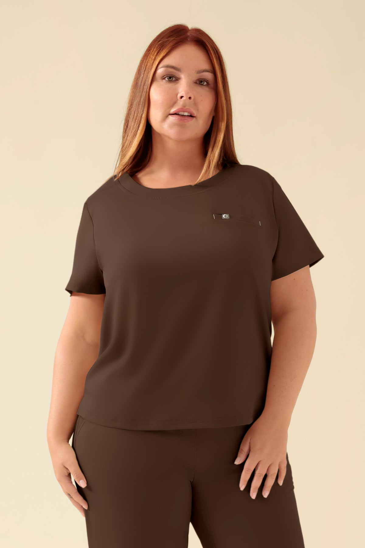 Eine Frau, die ein KAERE Shirt Damen – Rundhalsausschnitt in der Farbe Coffee trägt, ein dunkelbraunes, kurzärmeliges Oberteil mit Rundhalsausschnitt und einer kleinen Brusttasche., Vorderansicht Das Model ist 173 cm groß und trägt Größe XL