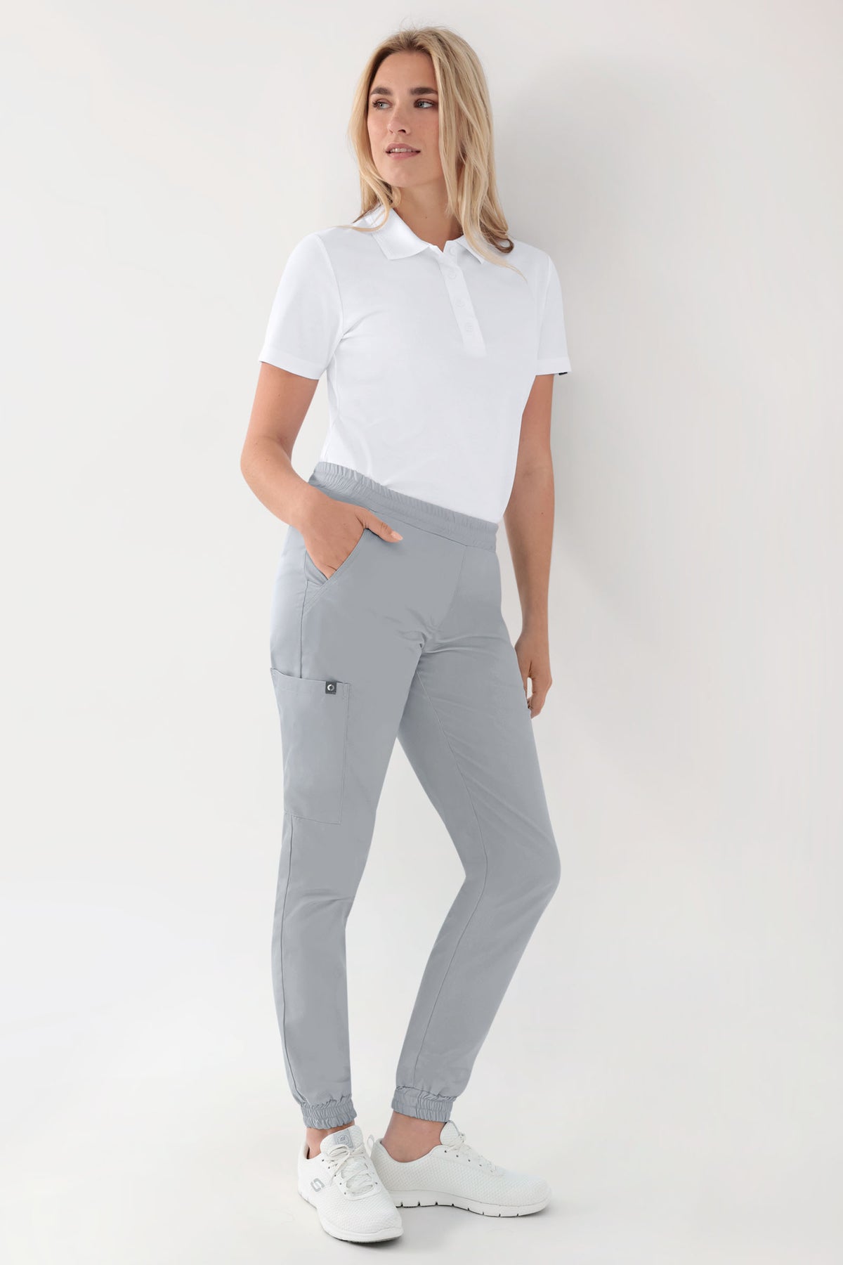 Eine Frau, die ein weißes Poloshirt, eine grüne Stretchhose von CLINIC DRESS im Jogginghosen-Stil in Perlgrau mit Cargotasche und elastischem Bund sowie weiße Sneaker trägt.