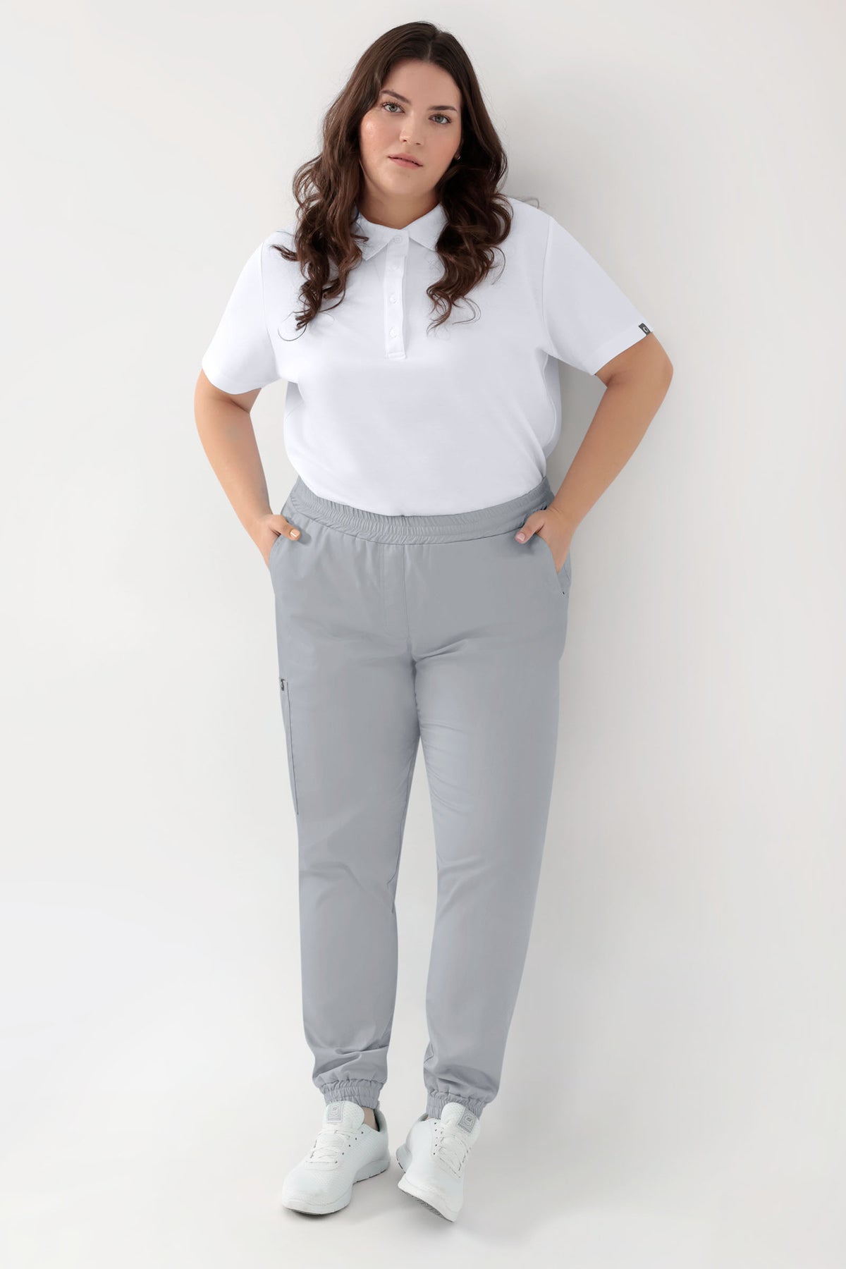 Eine Frau, die ein weißes Poloshirt, weiße Sneaker und eine Stretchhose CLINIC DRESS GREEN für Damen im Jogginghosen-Stil in Perlgrau mit bequemer, weit geschnittener Passform trägt., Vorderansicht 