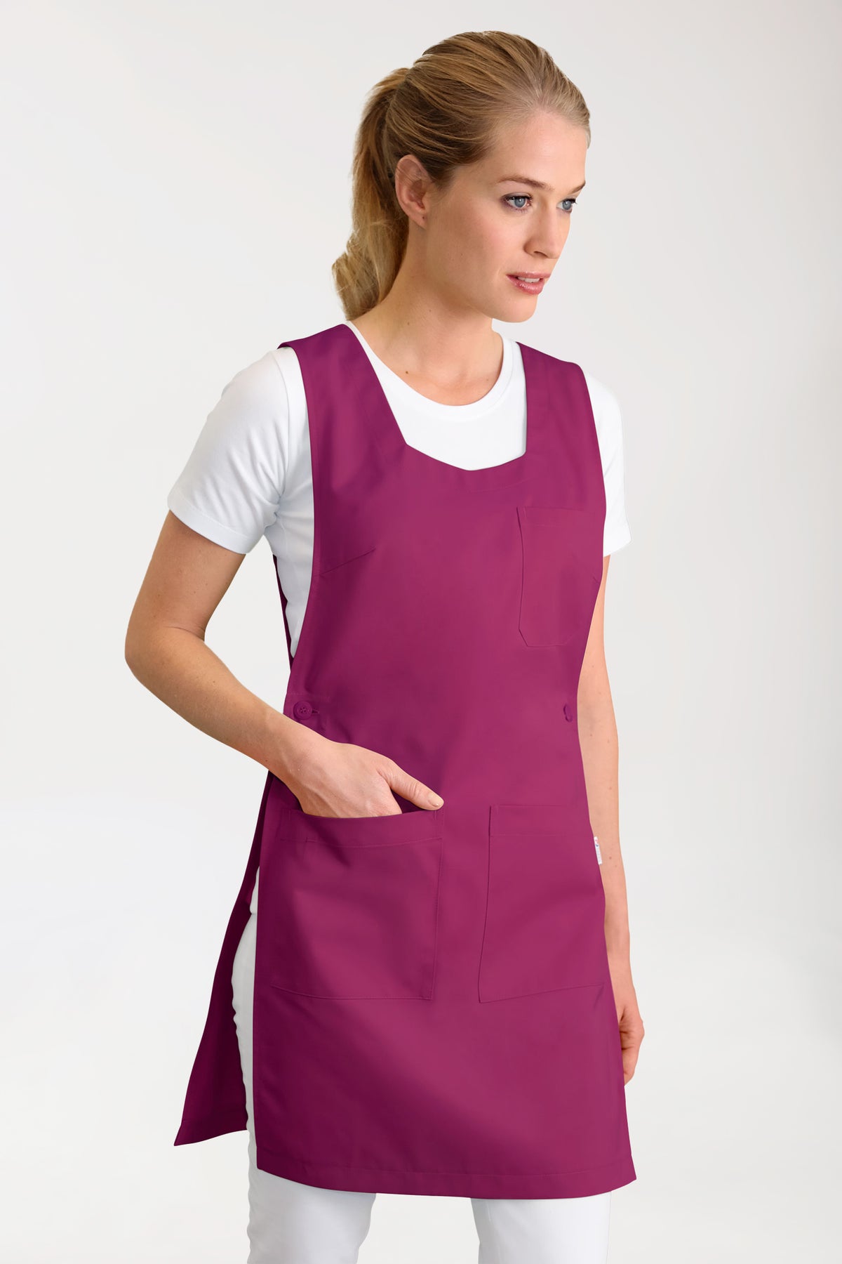 Eine Frau, die eine beerenfarbene CLINIC DRESS JUST STRONG Überwurfschürze für Damen mit Seitentaschen über einem weißen T-Shirt trägt.