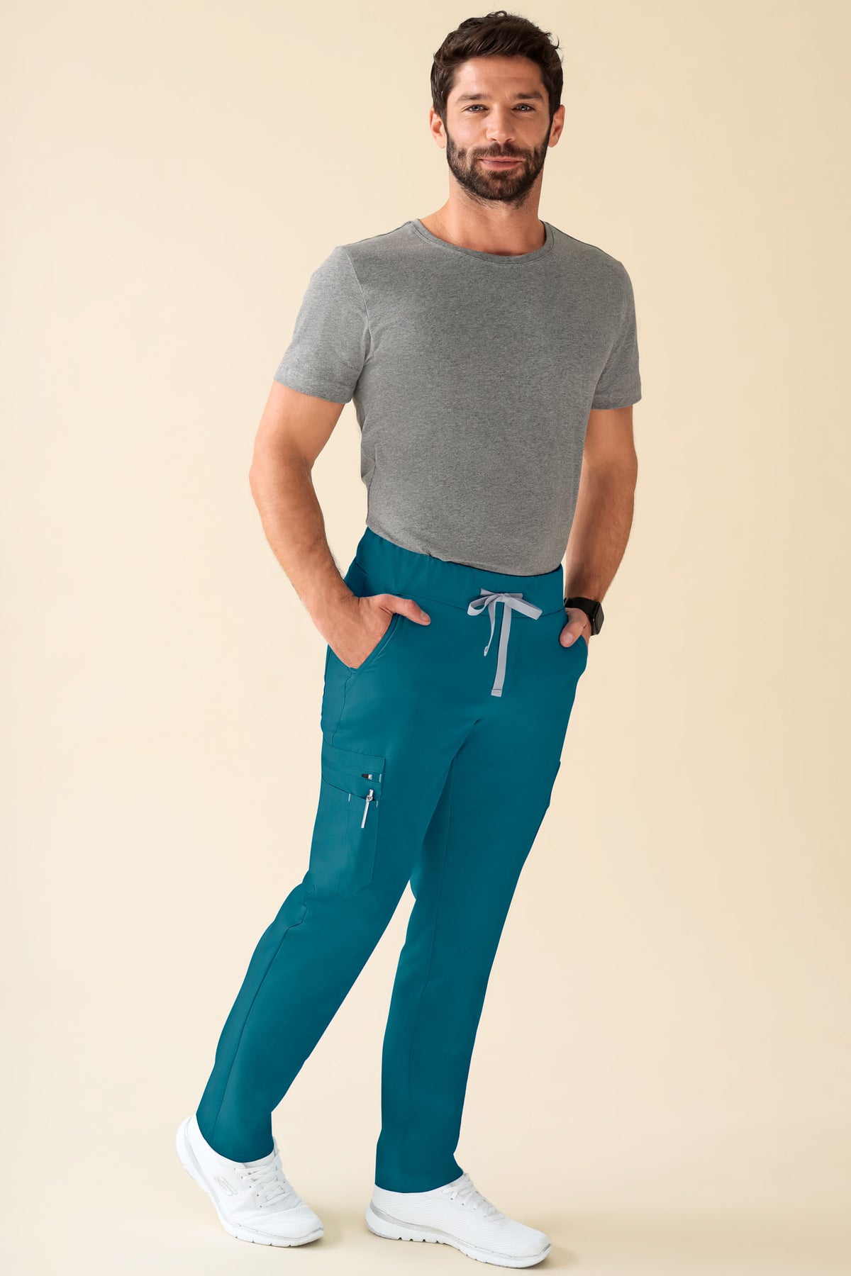 Ein Mann trägt ein graues T-Shirt und eine petrolfarbene KAERE-Herrenhose – gerader Saum, kurze Länge, mit Seitentaschen, Kordelzug in der Taille und kontrastfarbenen Riegelnähten.