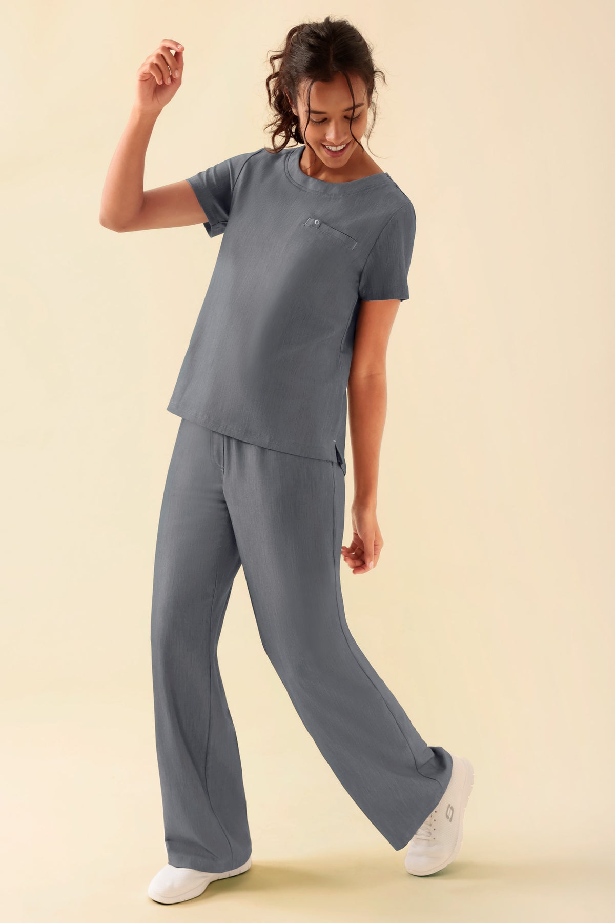 Eine Frau, die ein graues, meliertes KAERE-Shirt für Damen mit Rundhalsausschnitt und Brusttasche, dazu passende OP-Hosen und weiße Sneaker trägt.