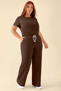 Eine Frau, die ein farblich passendes dunkelbraunes kurzärmeliges Oberteil und eine KAERE Hose Damen – weites Bein in der Farbe Coffee Scrub trägt – eine bequeme Hose mit weitem Bein und Kordelzug in der Taille.