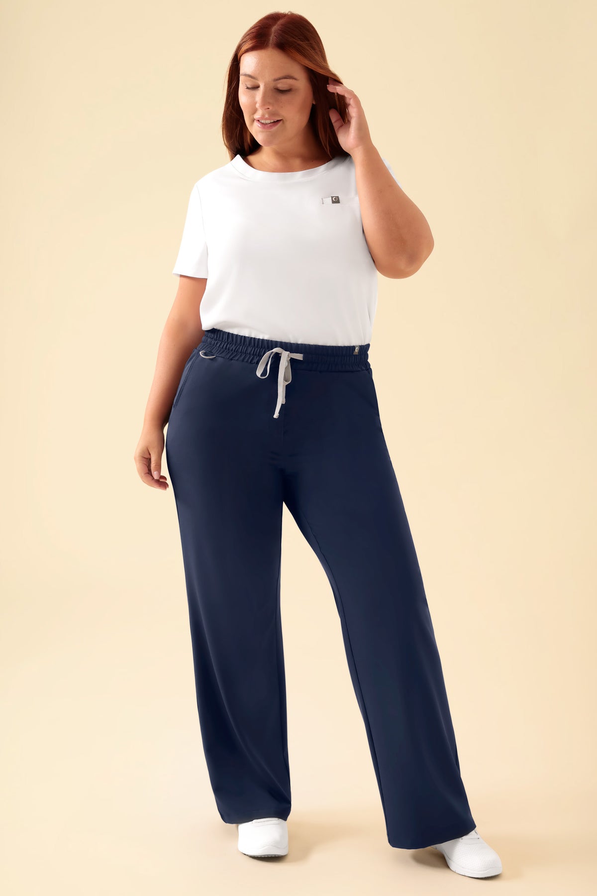 Eine Frau, die ein weißes T-Shirt und eine OP-Hose CLINIC DRESS KAERE Hose Damen – weites Bein in Marineblau trägt, die eine lässige, weit geschnittene Passform und einen Kordelzug in der Taille aufweist., Vorderansicht Das Model ist 173 cm groß und trägt Größe XL