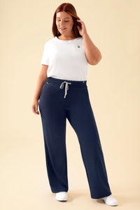 Eine Frau, die ein weißes T-Shirt und eine OP-Hose CLINIC DRESS KAERE Hose Damen – weites Bein in Marineblau trägt, die eine lässige, weit geschnittene Passform und einen Kordelzug in der Taille aufweist.