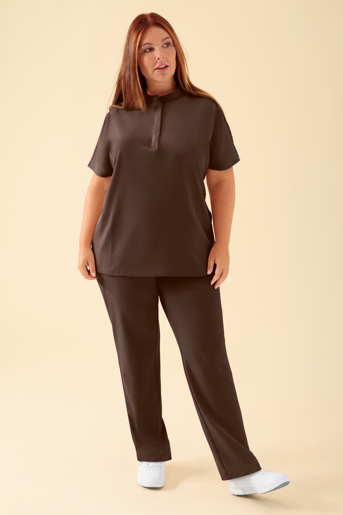 Eine Person, die einen CLINIC DRESS KAERE Schlupfkasack Damen – Stehkragen, Farbe: Kaffee trägt, der über einen Stehkragen, Seitentaschen und eine dazu passende Hose verfügt., Vorderansicht Das Model ist 173 cm groß und trägt Größe XL