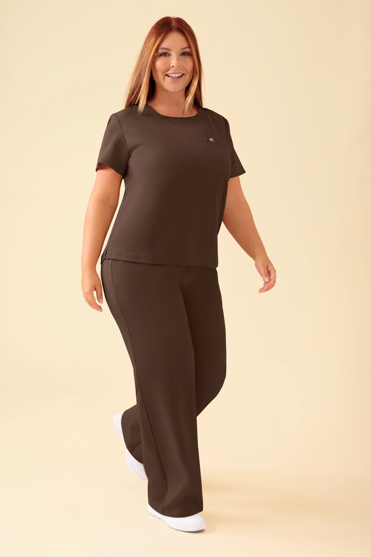 Eine Frau trägt ein braunes KAERE-Shirt für Damen – Coffee mit Rundhalsausschnitt und einer Brusttasche, dazu eine passende Hose und weiße Sneaker., Vorderansicht Das Model ist 173 cm groß und trägt Größe XL