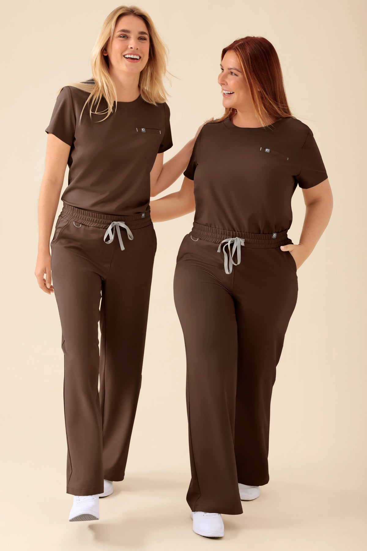 Zwei Frauen, die passende braune kurzärmelige OP-Oberteile und die KAERE Hose Damen – weites Bein in der Farbe Coffee mit einer bequemen, weit geschnittenen Passform und grauen Kordelzügen tragen., Vorderansicht 