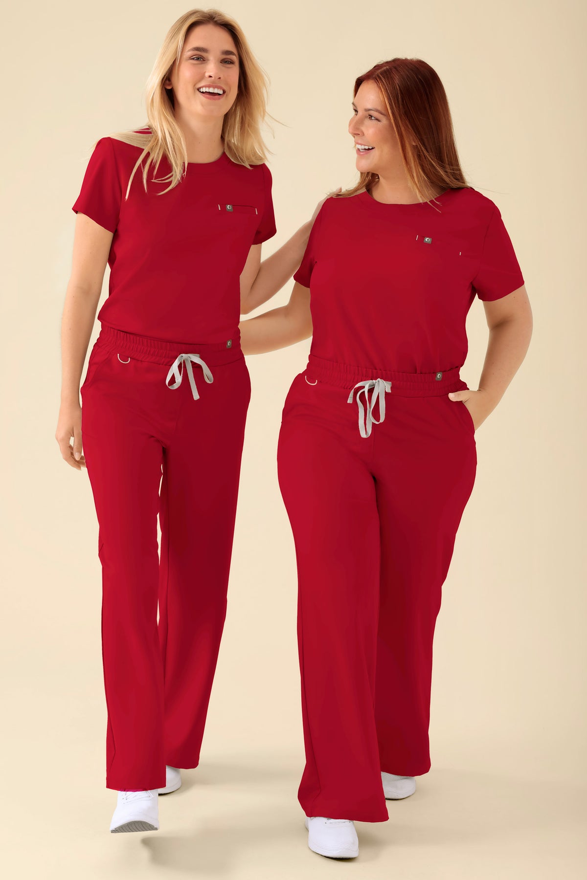 Zwei lächelnde Frauen, die passende rote KAERE-Hosen und -Oberteile tragen – rote Scrub-Oberteile und -Hosen mit weitem Bein und lässiger Passform sowie Kordelzug in der Taille., Vorderansicht 