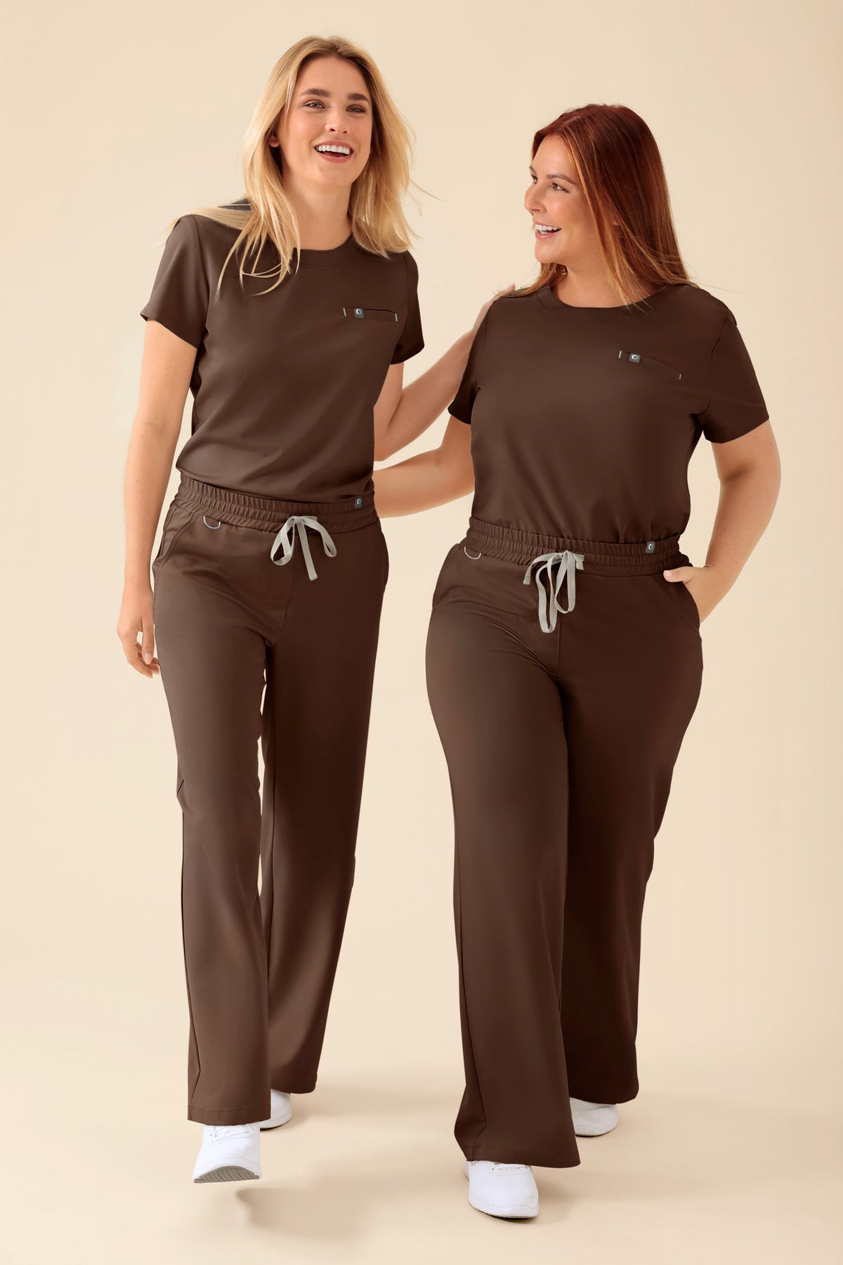 Zwei Frauen, die identische, kaffeefarbene KAERE Shirt Damen – Rundhalsausschnitt mit Rundhalsausschnitt und Brusttasche tragen, kombiniert mit OP-Hosen mit Kordelzug., Vorderansicht 