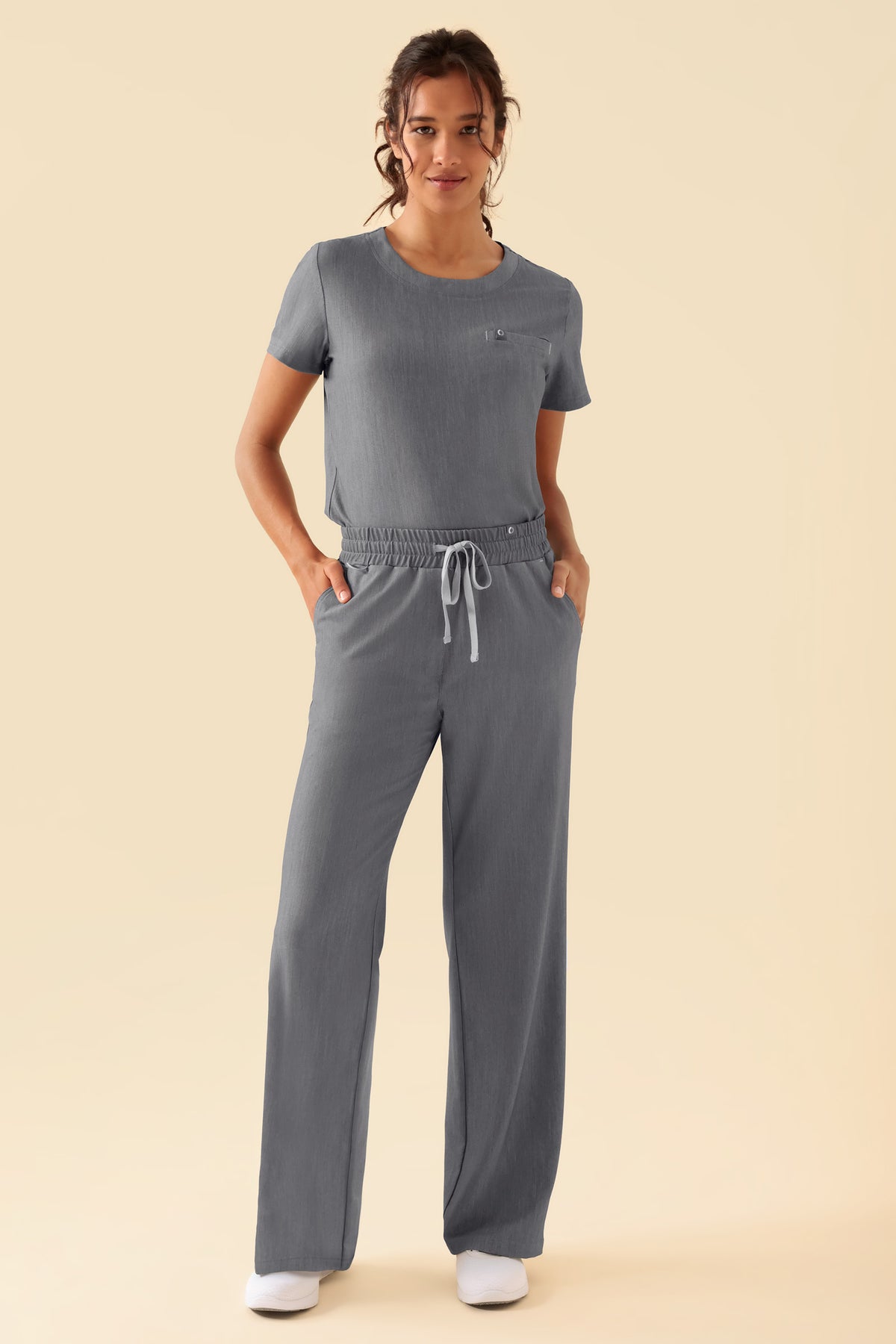 Eine Frau, die ein grau meliertes KAERE-Shirt für Damen mit Rundhalsausschnitt, Seitenschlitzen und einer Brusttasche trägt, dazu eine passende OP-Hose mit Kordelzug.
