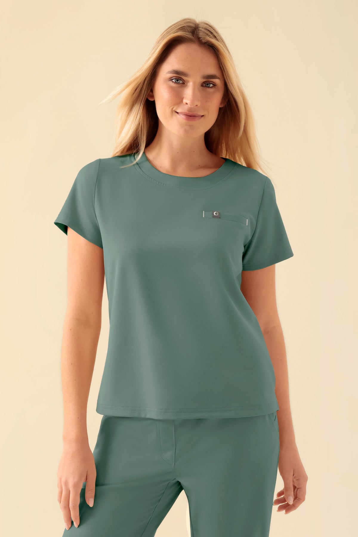Frau, die ein KAERE-Shirt Damen – Rundhalsausschnitt dunkelgrün trägt, ein dunkelgrünes OP-Oberteil mit kurzen Ärmeln, Rundhalsausschnitt, Seitenschlitzen und einer kleinen Brusttasche.