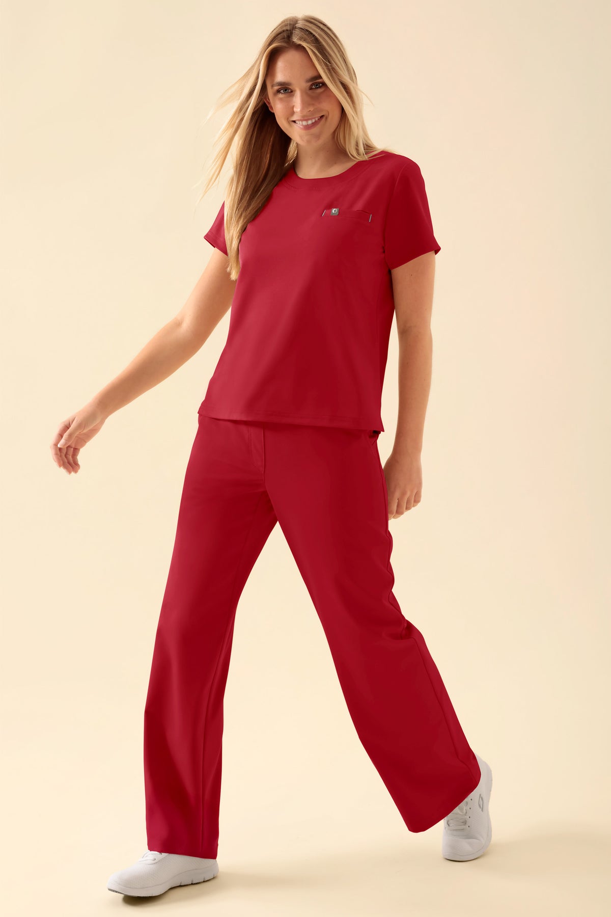 Eine Frau, die ein rotes KAERE-Shirt für Damen – Rundhalsausschnitt, rot – mit Rundhalsausschnitt und Brusttasche trägt, dazu eine passende rote OP-Hose und weiße Sneaker.